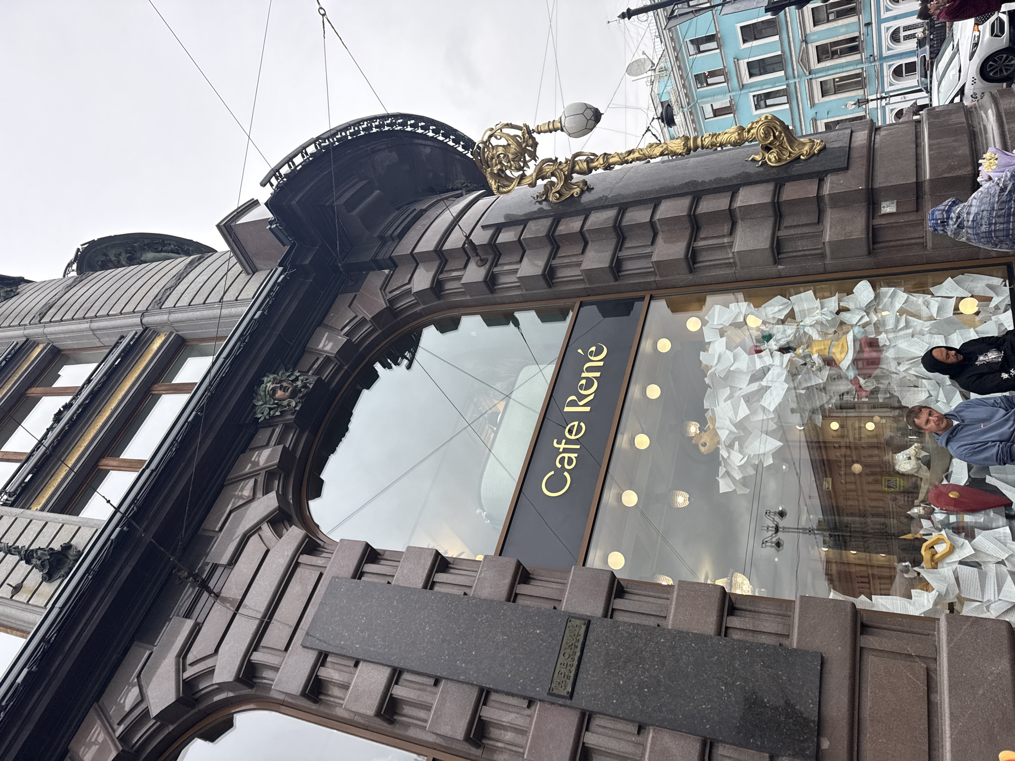 A low-angle, Dutch-angle shot shows the ornate facade of a building, likely a cafe or shop, with the text "Cafe René" visible on a dark sign with gold lettering. The building's exterior is made of dark, textured stone with intricate carvings and gold decorative elements, including a lamppost with a lion's head motif. Large windows reflect the cloudy sky and surrounding buildings. Inside the window display, a chaotic arrangement of crumpled white paper is visible, with some objects and a person partially obscured within.
In the reflection and visible within the display, a man in a blue hoodie appears to be looking towards the camera, possibly interacting with the photographer or observing the scene. Another person in a black hooded jacket is also partially visible. The sky is overcast, suggesting a cloudy day. The architecture suggests an urban setting, possibly European, with classical and art nouveau influences. The overall impression is one of grandeur and artistic display, with a touch of mystery due to the obscured elements in the window.