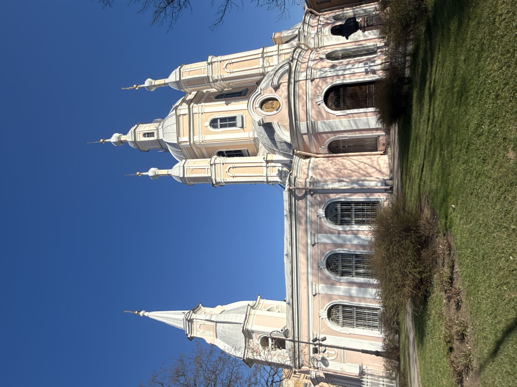 The Smolny Cathedral in Saint Petersburg, Russia, stands tall against a clear, blue sky. The cathedral's exterior is a blend of pink and white, featuring multiple domes and spires that reach upward. A patch of green grass runs along the right side of the frame, adjacent to the cathedral's walls, which are adorned with arched windows. A statue is visible on the building's facade. A few bare tree branches can be seen in the lower left corner.