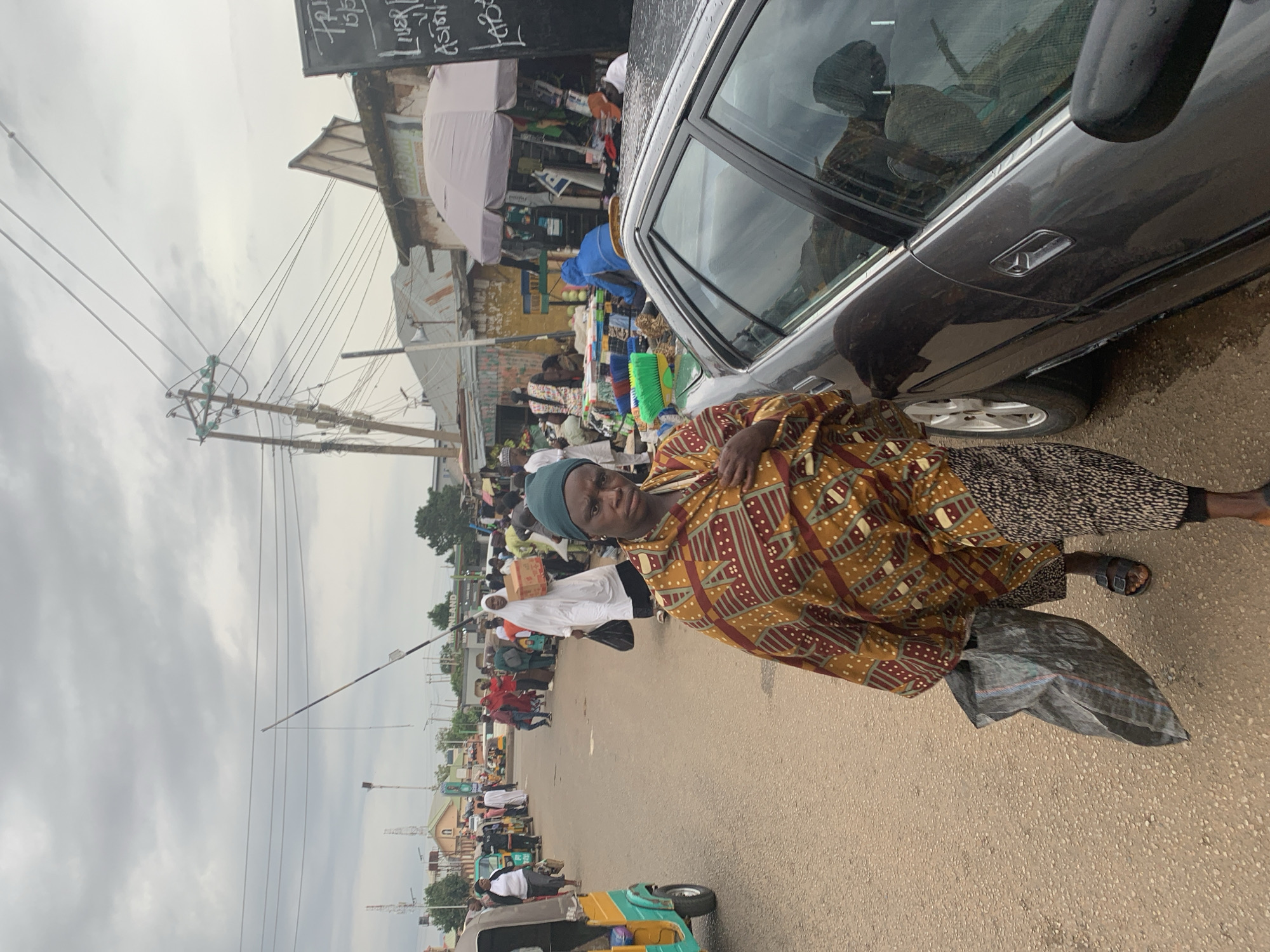 The image captures a street scene in Jos, Nigeria. An older woman, wearing a patterned brown and red top, a green head covering, and sandals, stands on the side of the road next to a gray car. She is holding a gray bag. Behind her, a busy street scene unfolds with people walking, and a small blue and yellow vehicle is in the lower left. In the background, there are market stalls and shops, suggesting a commercial area. Overhead are electrical power lines. The sky is overcast.