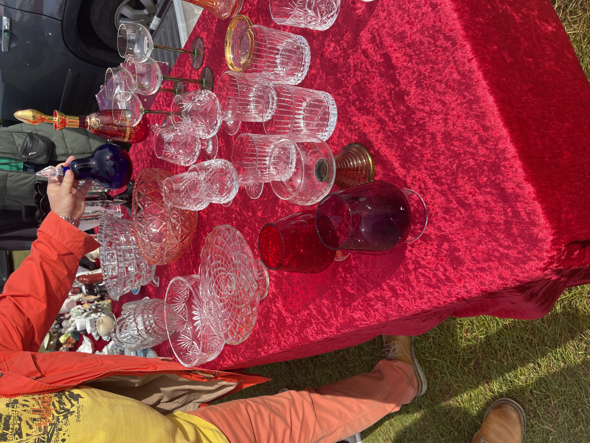 A person wearing an orange jacket and pants, and a yellow t-shirt, stands next to a red velvet-covered table with an array of glassware on display. The glassware includes clear crystal bowls, wine glasses, tumblers, and colored glass decanters in red, blue, and purple. Some of the items are being held up. A portion of a car is visible in the background, parked next to the table. The scene is set outdoors on grass. The location is suggested to be in Sevenoaks, United Kingdom.