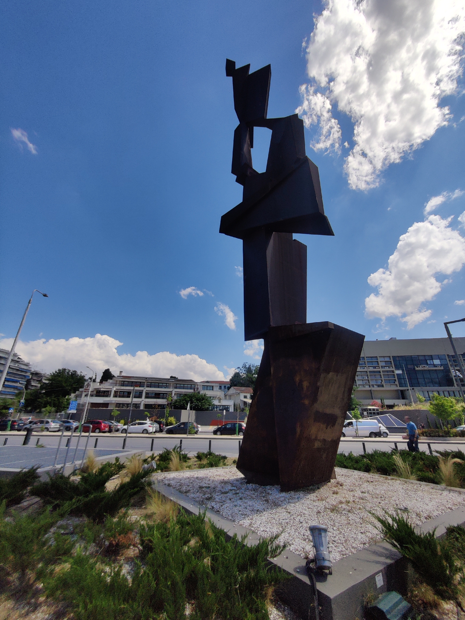 A tall, dark, abstract sculpture made of metal stands in the foreground of a street scene in Thessaloniki, Greece. The sculpture is angular and geometric, with sharp edges and planes, resembling a stylized human figure or an abstract form. It is situated on a raised circular platform covered in white gravel. Surrounding the platform is a bed of green bushes and ornamental grasses.
Behind the sculpture, a wide, multi-lane street is visible with several cars parked and driving. A tall street lamp with a single light fixture stands to the left of the sculpture. In the middle ground, there are several buildings, including a multi-story building with a light-colored facade and many windows, and a lower-lying building with a similar architectural style. Trees and greenery are interspersed between the buildings.
The background features a bright blue sky with scattered white, fluffy clouds. A person in a blue shirt is walking on the sidewalk to the right of the sculpture, and a white van is parked on the side of the street. The overall impression is a sunny day in an urban environment, with a striking piece of public art as the focal point.