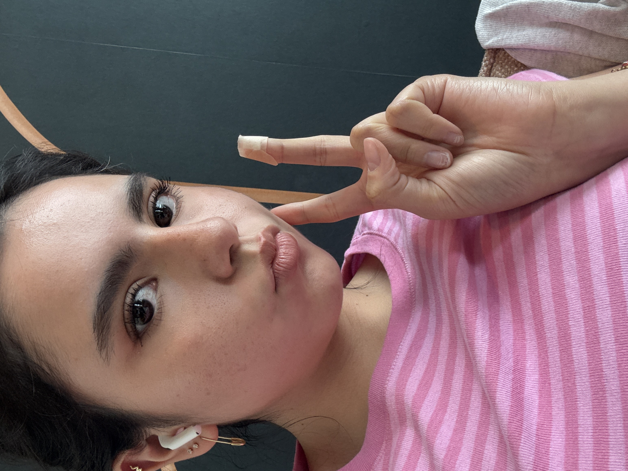A person with dark hair is facing the camera and making a peace sign with their right hand. The person is wearing a pink and white striped shirt. A bandage is on the index finger of the right hand. The person has brown eyes, dark eyebrows, and pink lips. An earbud and multiple earrings are visible in the person's left ear. The background is a dark gray color, with a thin wooden trim visible in the upper left corner. The photo appears to be taken indoors. It is not possible to identify if the picture was taken in Austin.