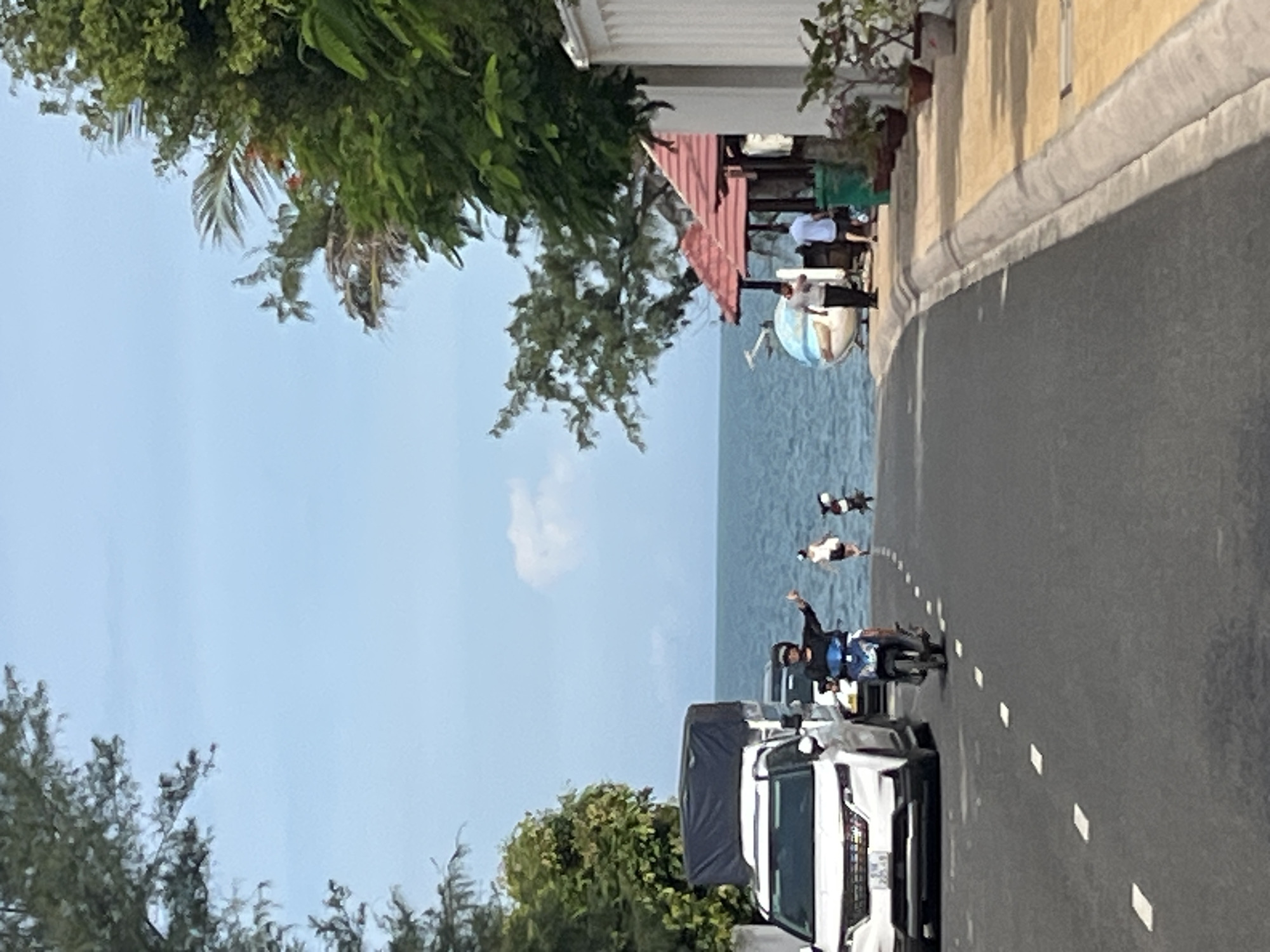 The image depicts a scene in Mũi Né, Vietnam, near the coastline. A paved road with white dashed lines runs towards the ocean. On the road, a white SUV is parked on the left, followed by a motorbike with a rider giving a thumbs-up gesture. Further down the road, two individuals are walking near the water's edge, and another motorbike is visible in the background. To the right of the road, a building with a red-tiled roof and open-air seating is situated near the beach, with several potted plants in front. The sea is calm and extends to the horizon under a blue sky with scattered clouds. Lush green trees and foliage frame the scene, with some vegetation also visible on the right side of the image. The overall atmosphere suggests a coastal town or resort area.