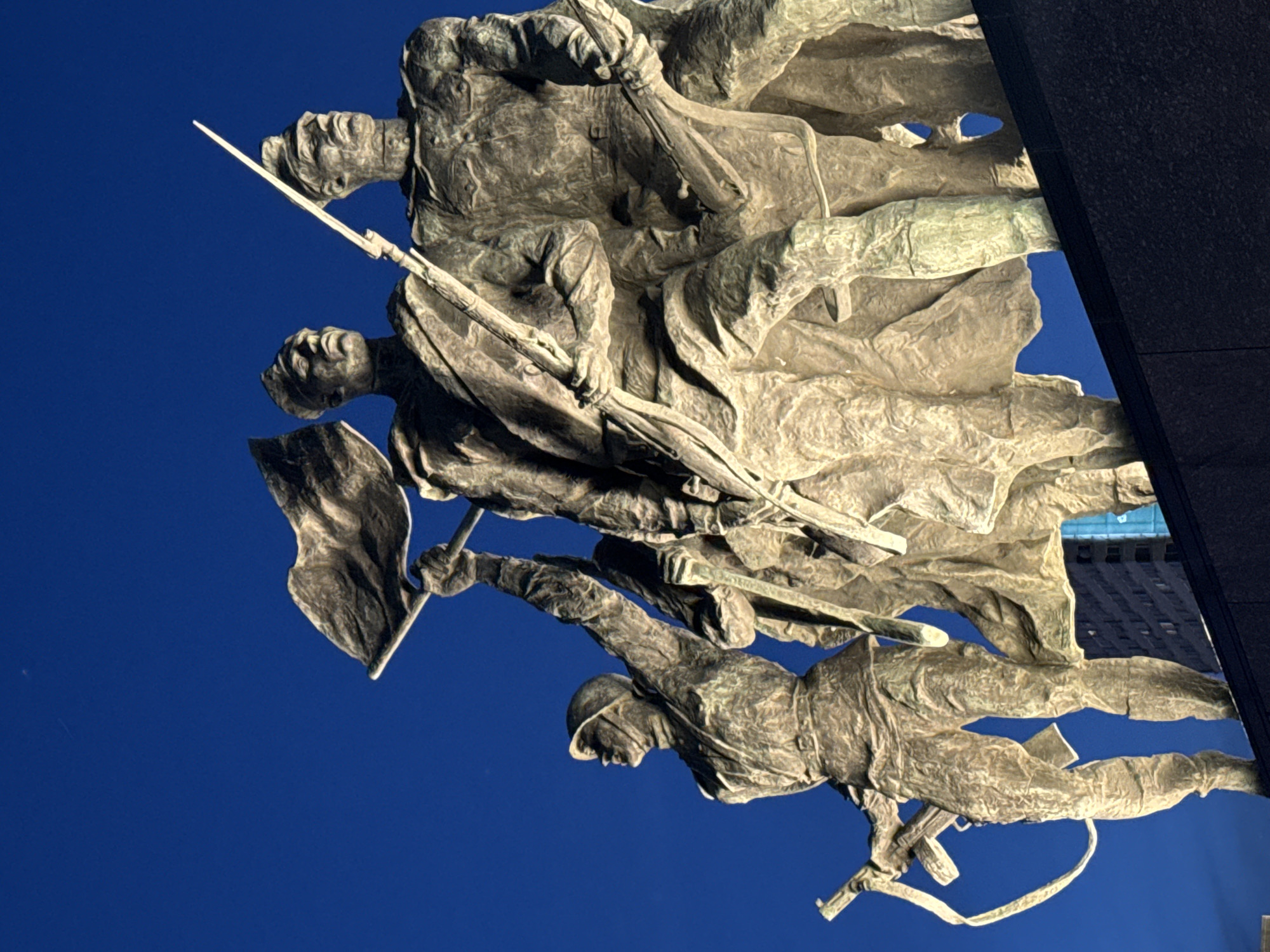 This image depicts a low-angle view of the Monument to the Heroic Defenders of Leningrad, located in Victory Square, Saint Petersburg, Russia. The monument comprises a large bronze sculptural group featuring at least five Soviet soldiers in dynamic postures, illuminated from below against a deep blue night sky. The figures are rendered in patinated bronze, showcasing detailed military uniforms from the mid-20th century. The leftmost soldier wears a helmet, carries a rifle with a sling, and gazes upward. Adjacent, a soldier holds a large flag aloft. The central figure, oriented forward, holds a rifle equipped with a fixed bayonet. A fourth soldier to the right also carries a rifle, facing forward. The statues exhibit significant textural detail and strong contrasts due to the direct upward illumination. The base upon which the statues stand is dark and textured. In the lower-right background, a segment of a modern high-rise building with illuminated windows is visible.