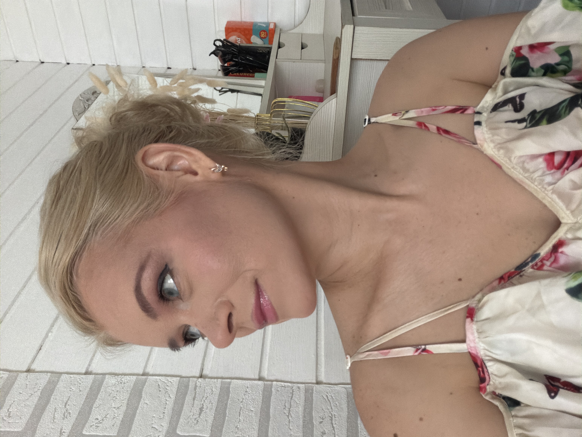 This is a close-up, sideways portrait of a young woman with blonde hair, styled in a messy bun. She is wearing a floral-patterned top with thin straps. Her makeup includes eyeshadow and lip gloss. Her left ear is visible, adorned with a small earring. The background consists of white, vertically paneled walls, with a glimpse of a shelf unit holding various objects, including what appears to be tissue boxes and other toiletries. The overall lighting suggests an indoor setting, possibly during the daytime. The image has a soft, slightly grainy texture.