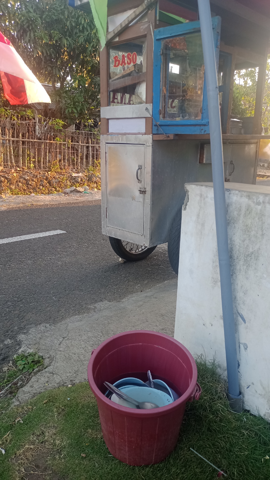 A mobile food cart, labeled "BASO," is parked on the side of a road in Pakemitan, Indonesia. The cart is constructed of wood and metal, with a glass display case showcasing food items. A large red bucket filled with water and dishes sits on the grassy shoulder of the road, to the right of the cart. The road has a white stripe marking its edge. Behind the cart, a bamboo fence separates the road from a patch of dry earth and vegetation. Trees with green foliage are visible in the background. The scene appears to be illuminated by late afternoon sunlight.
