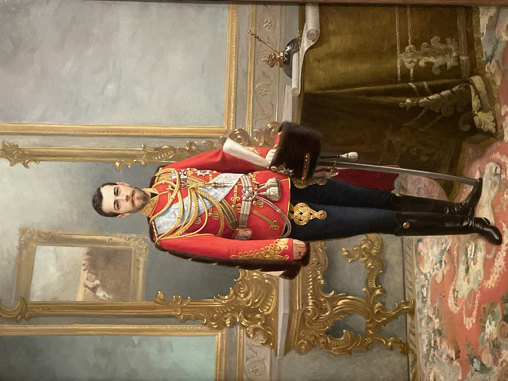 The image is a portrait painting depicting a man in a highly ornate red military uniform with gold embellishments, dark blue trousers, and black boots. He has a beard and is wearing numerous medals and sashes. A black fur hat rests on a table next to him, along with a sheathed sword. He stands on a patterned carpet in a room with pale blue walls. A gilded frame surrounds a painting behind him on the left. To the right, there is a partially visible dark green curtain or drapery with gold trim. The location is likely a regal or official setting, possibly a state building or palace, in Saint Petersburg, Russia, given the style and detail of the painting and the nature of the man's attire.