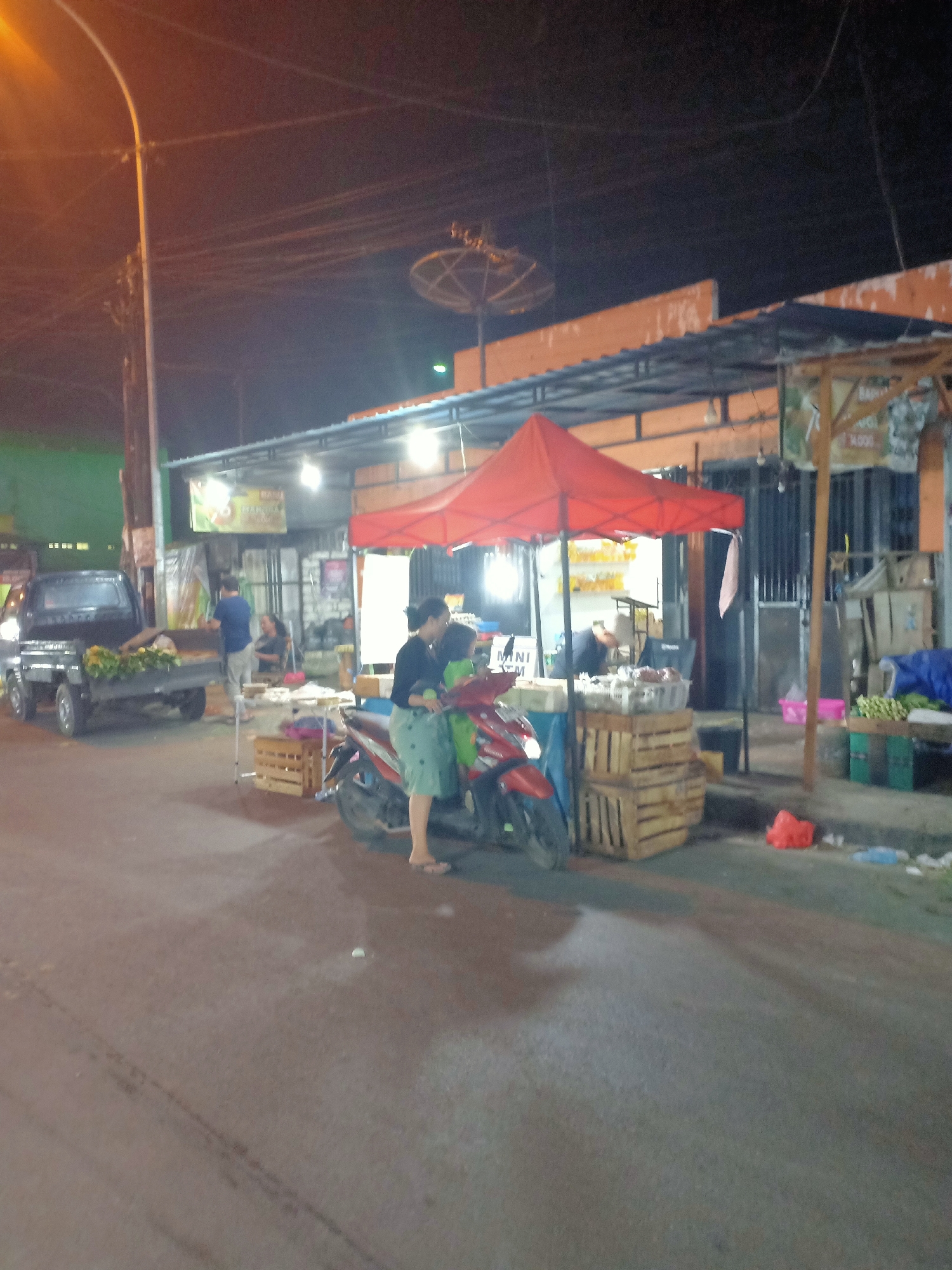 This is a night-time street scene in Semanding, Indonesia, likely a market area. In the foreground, a red motorcycle is parked, with a woman in a black top and light-colored shorts standing next to it, appearing to interact with someone behind the motorcycle or the market stall. Behind her, a market stall is set up under a red canopy, with crates of produce and other goods visible. Further to the left, a black pickup truck is parked, loaded with what appears to be produce. The area is illuminated by streetlights and lights from the market stalls, creating a lively but somewhat dimly lit atmosphere. In the background, a satellite dish is visible on top of a building, and other market stalls or shops line the street.