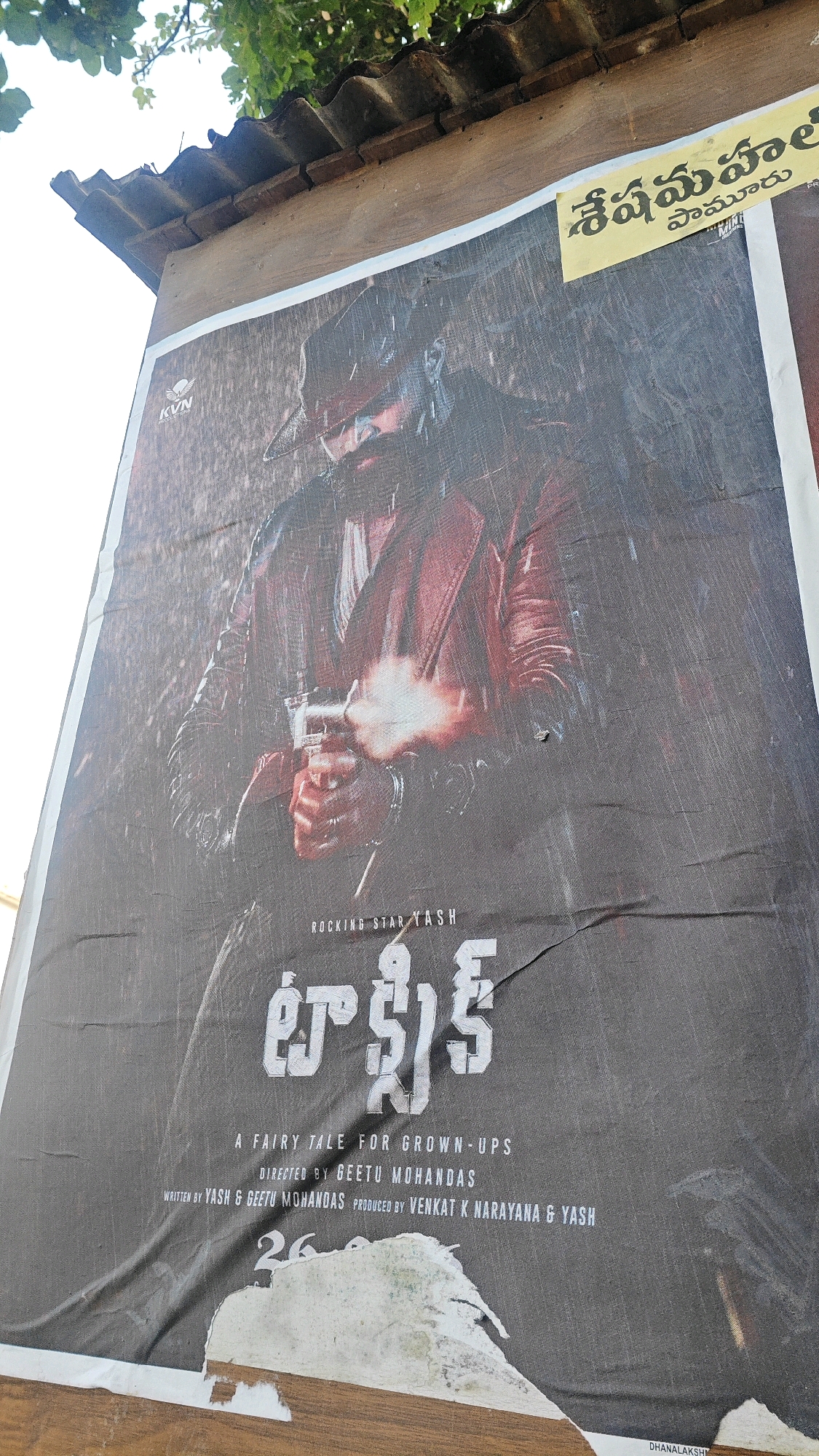 A movie poster, featuring actor Yash, is plastered on a wooden structure under a tiled roof. The poster advertises the film "Toxic" and is in the Telugu language, with English text below. The actor is depicted holding a gun, with smoke emanating from the barrel. The poster is weathered and torn in places, with a portion of it peeling away at the bottom. A yellow banner at the top of the poster displays text in Telugu script. The overall scene suggests an outdoor setting, likely in a public area where movie posters are displayed. The provided context indicates this scene is located in Pamur, India.