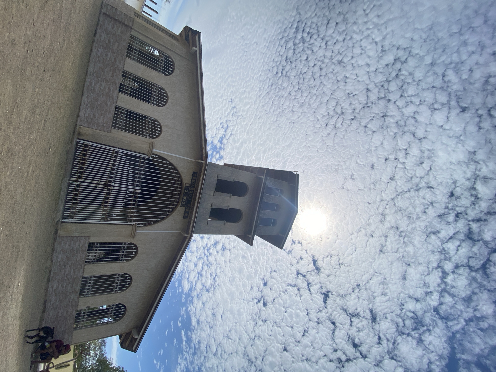 The image shows a low-angle view of a church facade against a backdrop of a sky densely populated with altocumulus clouds, partially obscuring the sun. The church is a light beige color and features arched windows with metal grilles. A prominent tower-like structure with open archways rises from the main building. The ground adjacent to the church is visible and appears to be a mixture of dirt and gravel. In the lower left corner, there are partial figures of individuals and possibly a dog. The location is identified as Juan Griego, Venezuela.