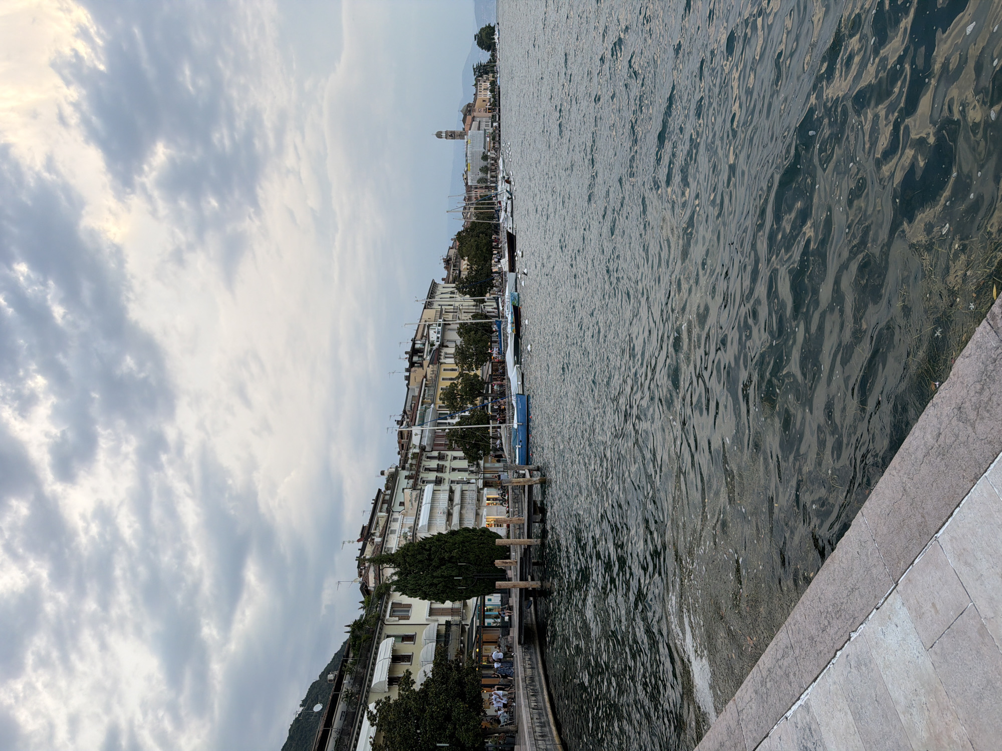 The image shows a waterfront scene in Salò, Italy, under a cloudy sky. In the foreground, a stone promenade runs along the left side of the frame, with grey and beige paving stones. The water of Lake Garda is to the right, exhibiting dark blue-green hues with ripples and some scattered debris on the surface.
Along the waterfront is a row of buildings with multi-story facades in pastel colors, featuring awnings, balconies, and arched windows. Trees are interspersed between the buildings and along the promenade. Several boats are moored or sailing in the lake, including sailboats and a small blue vessel. Some people are visible on the promenade, seated at outdoor cafe tables. A wooden pier extends into the water from the promenade, with several upright pilings. In the distance, a bell tower rises above the buildings, characteristic of an Italian town. The overall atmosphere is calm and picturesque.