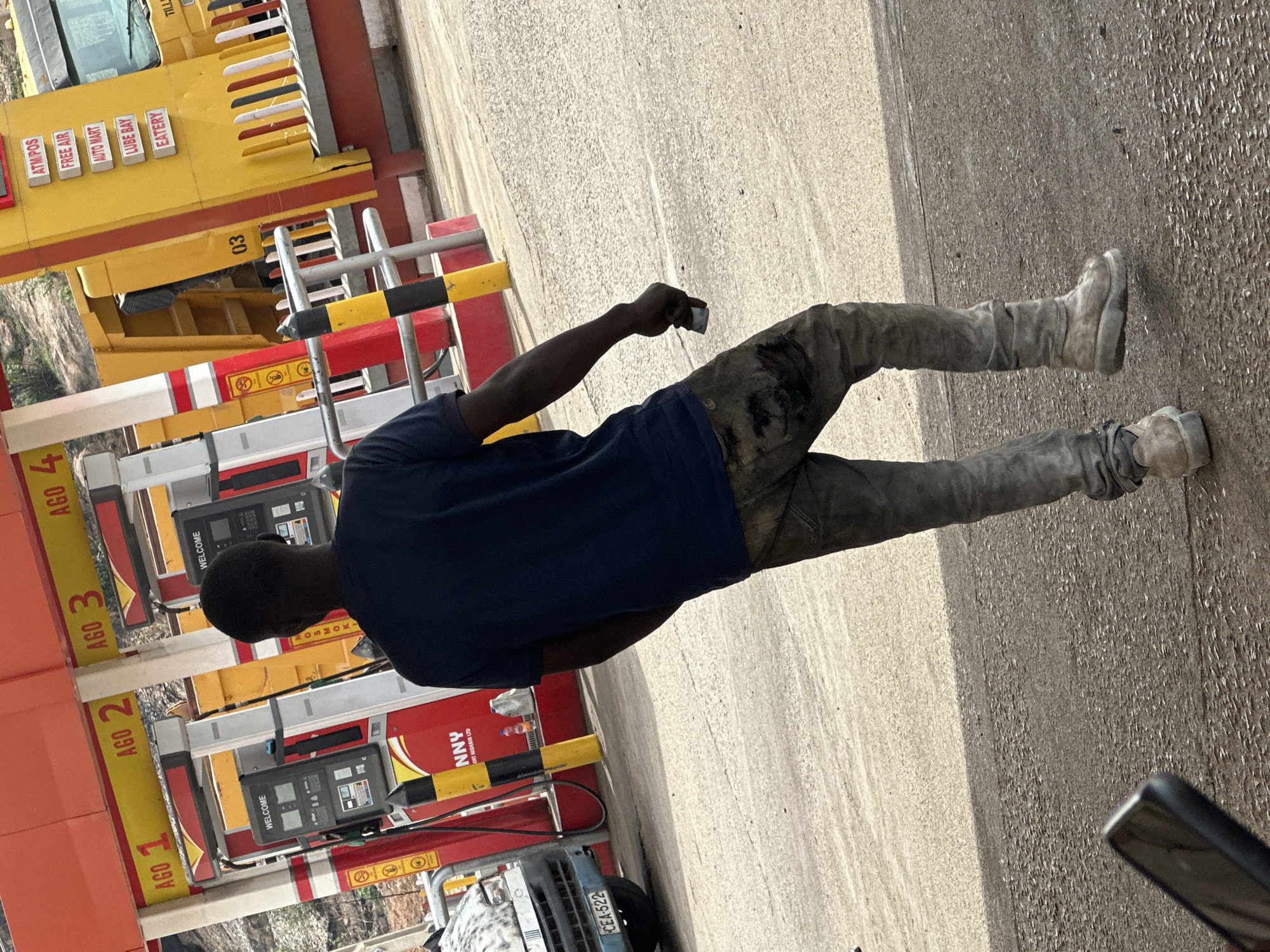 The image depicts an outdoor scene at a gas station or fueling station in Gura Top, Nigeria, during the daytime under bright sunlight. The central subject is a person, likely male, with dark skin, viewed from behind. He is wearing a dark blue short-sleeved t-shirt and light-colored pants or jeans that show signs of dirt or stains, especially around the thighs and knees. His light-colored boots or shoes also appear worn and soiled. He holds a small, dark rectangular object, possibly a phone, in his right hand.
In the background, a row of red and yellow fuel pumps or dispensers is visible. Each pump has a black screen displaying "WELCOME" and is labeled with numbers such as "AGO 1", "AGO 2", "AGO 3", and "AGO 4", likely referring to Automotive Gas Oil pumps. One pump base includes the text "NNY ENT NIGEAIR LTD." Above the pumps, a yellow building or canopy features signs for various services: "ATM/POS," "FREE AIR," "AUTO MART," "LUBE BAY," and "EATERY," along with the number "03." A partially visible "NO SMOKING" sign is also present.
Part of a blue vehicle with the license plate "CEA-522" can be seen near the fuel pumps. The ground is a light-colored, paved surface, illuminated by strong daylight, creating distinct shadows. The overall scene suggests a busy service station environment.