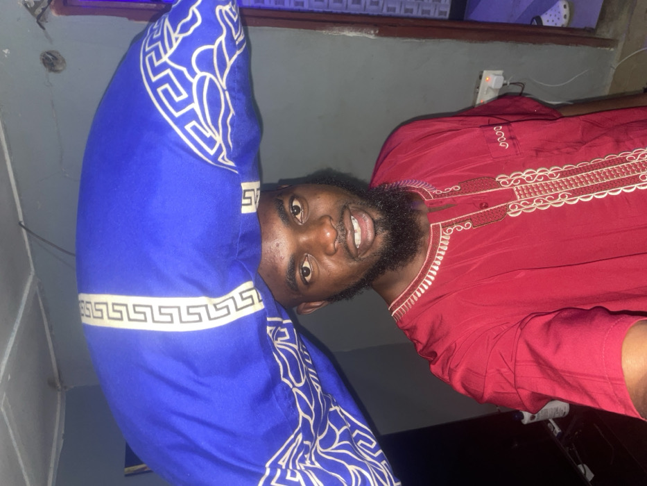A man with a beard smiles at the camera while holding a blue patterned pillow. He is wearing a maroon shirt with a decorative white collar. The background includes a wall with an electrical outlet and what appears to be a light fixture or part of a shoe. The image is slightly angled.