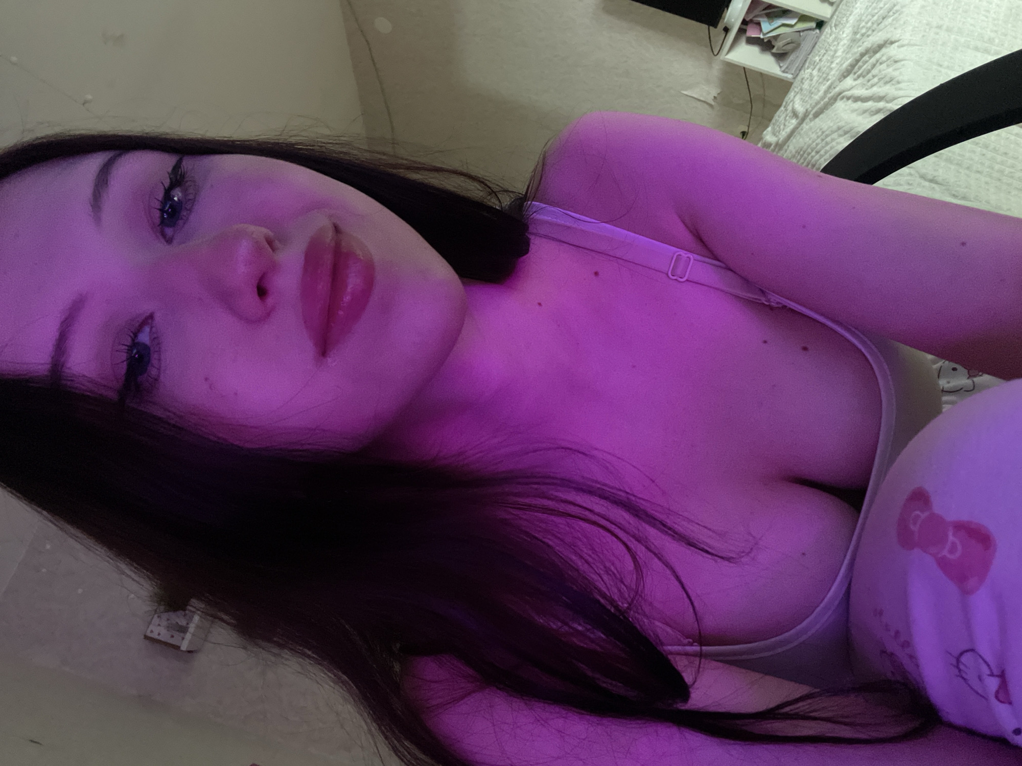 The image presents a young woman in an indoor setting, identified as a private residence in Votkinsk, Russia. The scene is illuminated by a prominent magenta light source, bathing the subject and portions of the background in a deep purple hue. The woman, with long dark hair, looks directly towards the camera. She wears a light-colored bra or camisole, exposing cleavage, and light-colored pajama bottoms featuring a cartoon character pattern on her upper left thigh. Her facial features include dark, glossy lipstick and defined eye makeup. The background on the right includes a white quilted bed, a partially visible black chair, and a white shelving unit containing various items. A small, decorative object is fixed to the wall on the left, and electrical wiring is discernible on the wall behind the bed.