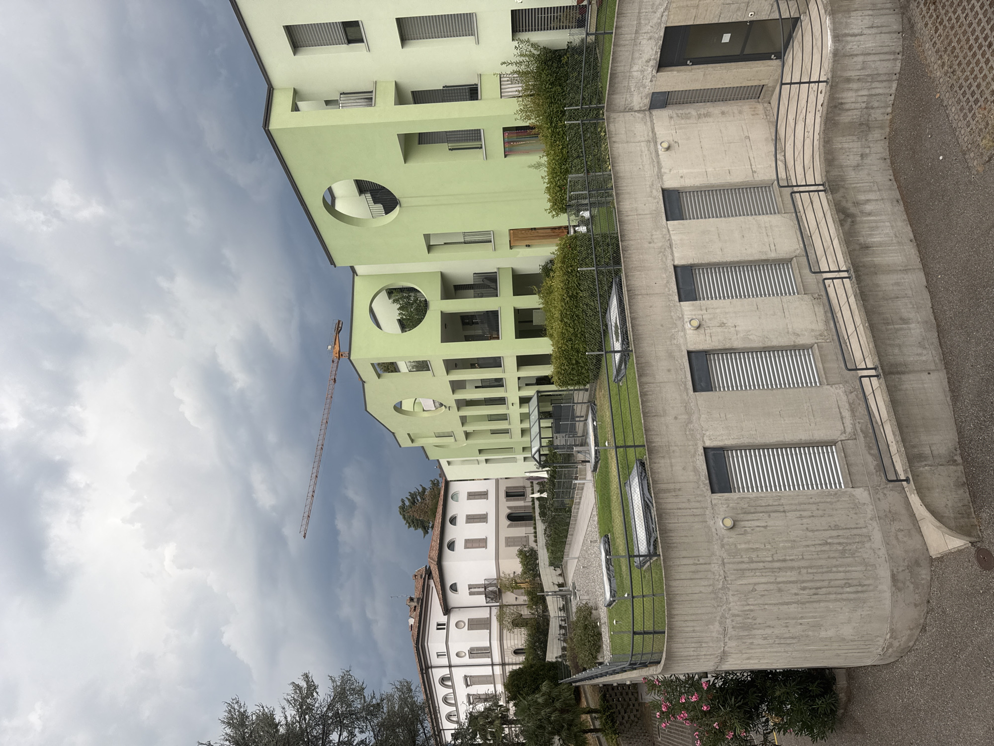 The image depicts a residential area in Balerna, Switzerland, on a cloudy day. In the foreground, a concrete structure with roller shutters and a curved ramp leads to an entrance. Above this, a rooftop garden with two covered rectangular structures is visible, bordered by a metal fence.
Further back, a distinct modern apartment building with a light green facade dominates the scene. This building features large circular cutouts on its upper floors, revealing balconies and internal staircases. Next to it, a more traditional white building with arched windows and shutters stands.
In the background, a construction crane is visible against the overcast sky, suggesting ongoing development in the vicinity. The landscape is dotted with trees and shrubs, including a flowering bush with pink blossoms on the left side of the frame. The overall impression is one of a well-maintained, possibly upscale, residential and developing area.