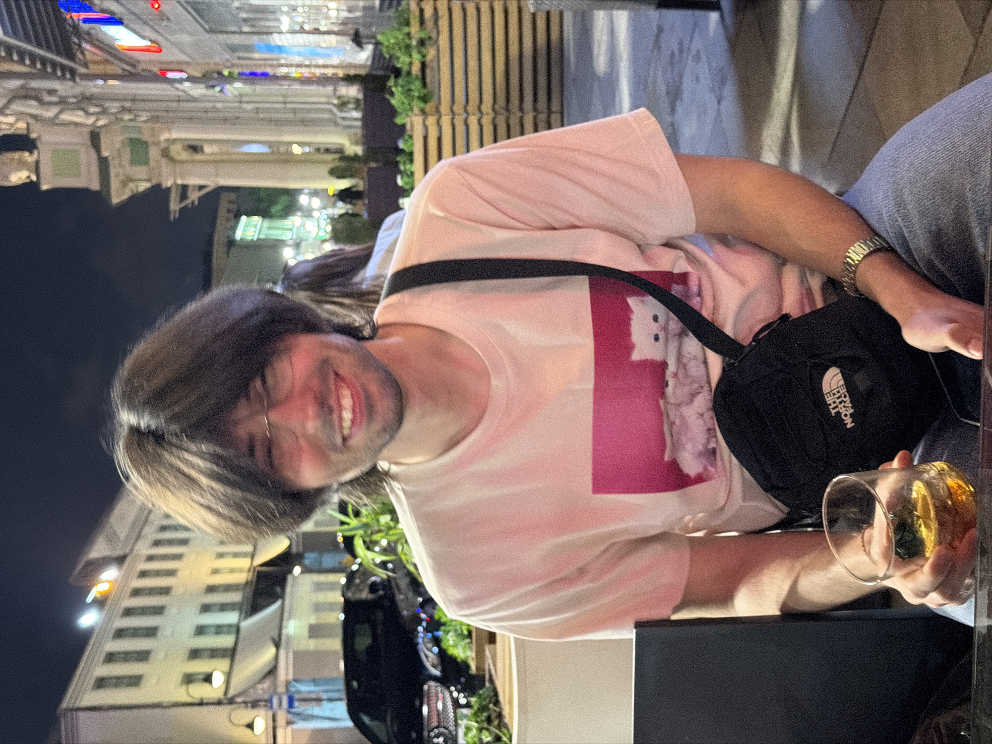 A medium shot depicts a man with brown hair and a slight beard, wearing glasses and a light pink t-shirt with a graphic. He is smiling and holding a glass of amber-colored liquid. A black shoulder bag with "The North Face" logo is visible on his shoulder. The backdrop shows an urban landscape, suggesting Moscow, Russia, based on the architecture and street lighting. Buildings with multiple stories are seen in the background, along with streetlights and traffic. A car is parked nearby. The setting implies an outdoor cafe or a street-side environment at night.