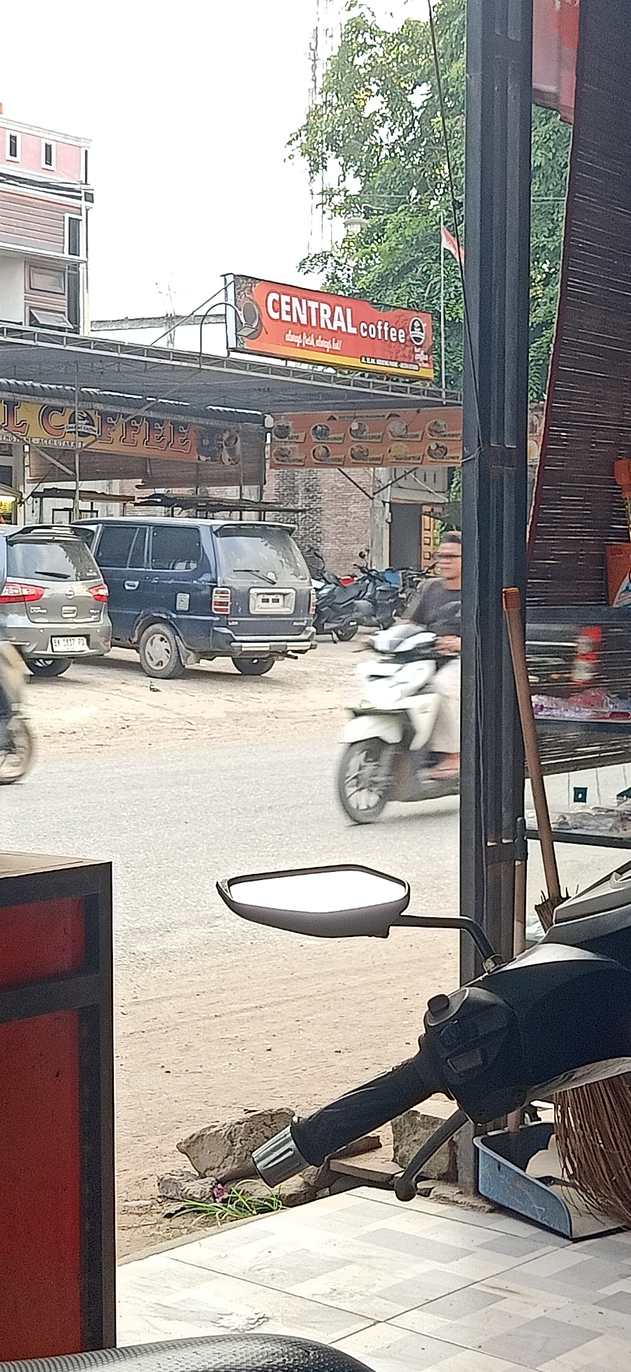 The image depicts a street scene in Gampongteungoh, Indonesia, likely in front of a commercial establishment. A prominent red and white banner for "CENTRAL COFFEE" is visible, with smaller text below reading "always fresh, always hot!". To the left, a pink and white building with horizontal siding stands. The foreground is dominated by a motorcycle handlebar and side mirror.
In the middle ground, a dark blue minivan is parked on a dirt or gravel surface. To its left, the rear of a silver hatchback car is visible, displaying a license plate that appears to read "EK 2837 FD". A white scooter with a rider, whose face is blurred, is moving from right to left across the road. Further in the background, several other motorcycles are parked. The environment appears to be a somewhat dusty, outdoor setting with trees and a brick building visible behind the coffee shop banner. A dark vertical pole, possibly part of a structure, is in the right foreground, with a bamboo-like material attached to it.