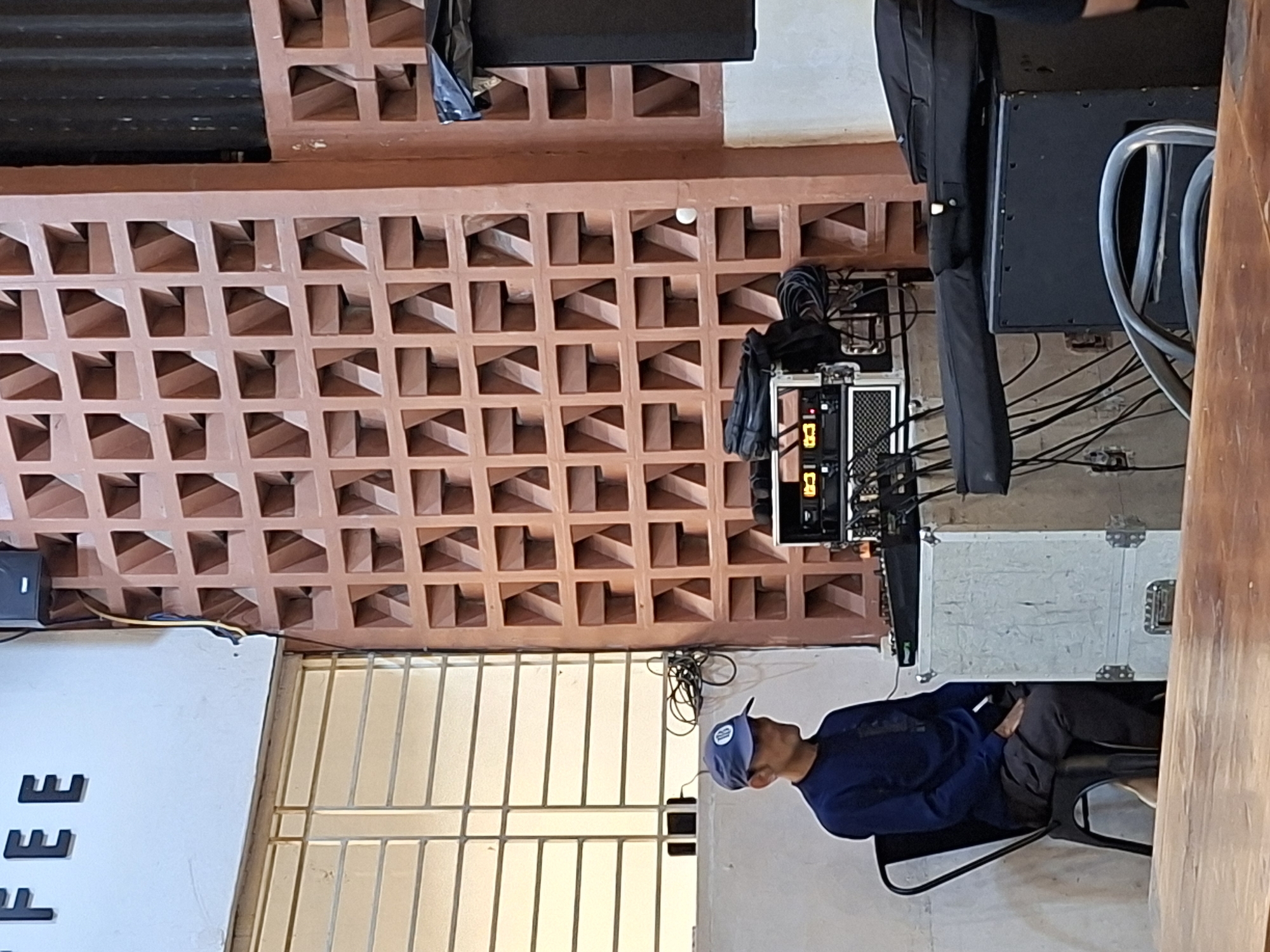 A medium, upward shot captures a man seated in a chair within a room, likely a cafe, in Lhokseumawe, Indonesia. He wears a blue long-sleeved shirt, gray pants, and a blue baseball cap. His body is oriented towards the right.
Behind him, a metal and white wire gate is visible. Above the gate, a white sign displays what appears to be the letters "FEE" in a vertical orientation.
The upper portion of the frame showcases a coral-colored brick wall featuring an intricate pattern of squares with diagonal openings. Directly behind the man's head, electronic equipment can be seen; it is on a silver rack, and is surrounded by black cables and wires. Additional sound or performance equipment is visible to the right, stacked and cased. The overall impression is that this is a stage or performance area.
