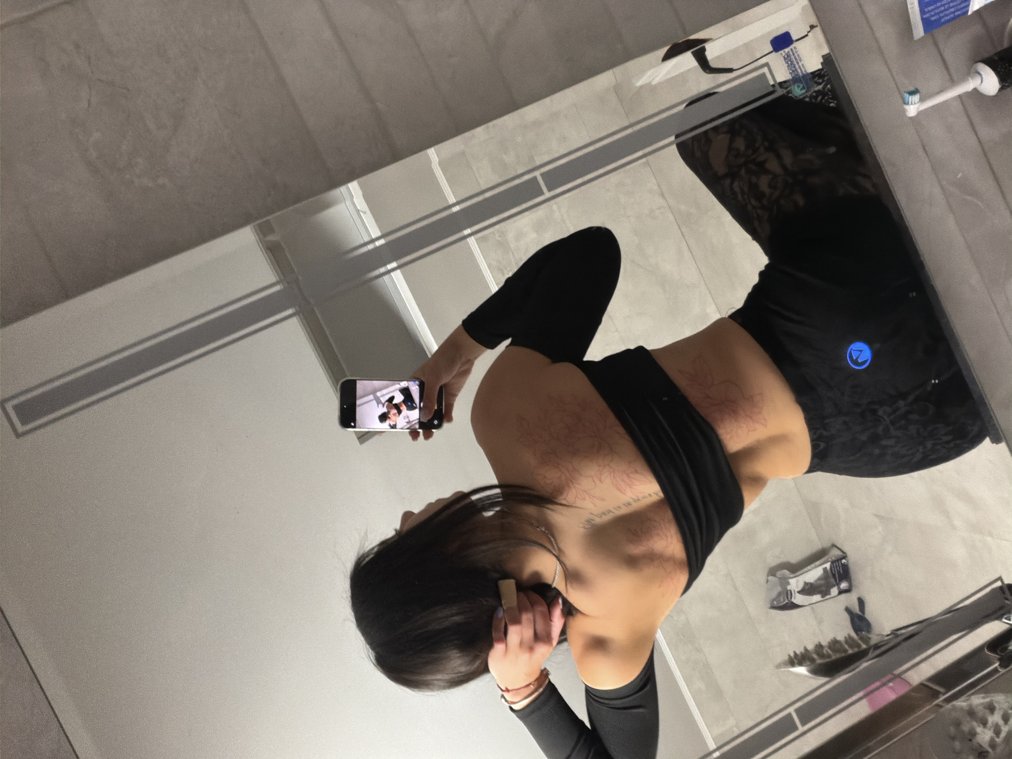 A woman is photographing her reflection in a mirror, likely in a bathroom setting. The setting includes elements associated with a bathroom, such as tiling and a toothbrush. The location is not apparent from this image.