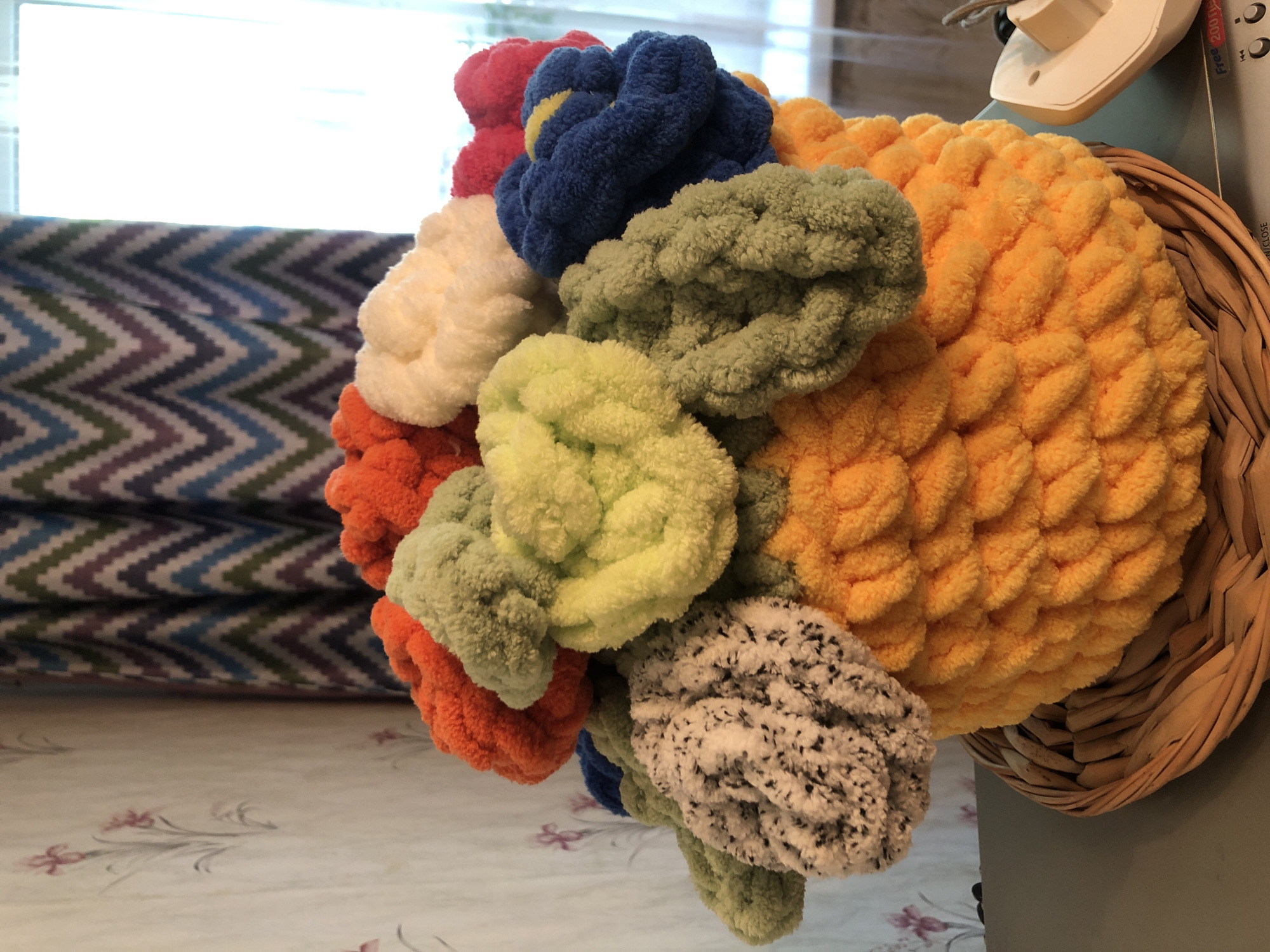 A close-up, eye-level shot shows a woven basket filled with colorful, fluffy yarn flowers. The basket is made of natural fibers and has a textured weave. The yarn flowers are in various bright colors including orange, yellow, green, white, blue, and red, with one flower having a black and white speckled pattern. The flowers are densely packed and appear to be made from chunky, loop yarn. The background consists of a patterned curtain with zig-zag stripes in shades of purple, green, and blue, and a wall with a floral wallpaper pattern featuring pink flowers and green stems. A window is visible behind the curtain, letting in bright natural light. A beige electrical device is partially visible on the right side of the frame. The scene appears to be an indoor setting, possibly a home, with no indication of a specific city or country.