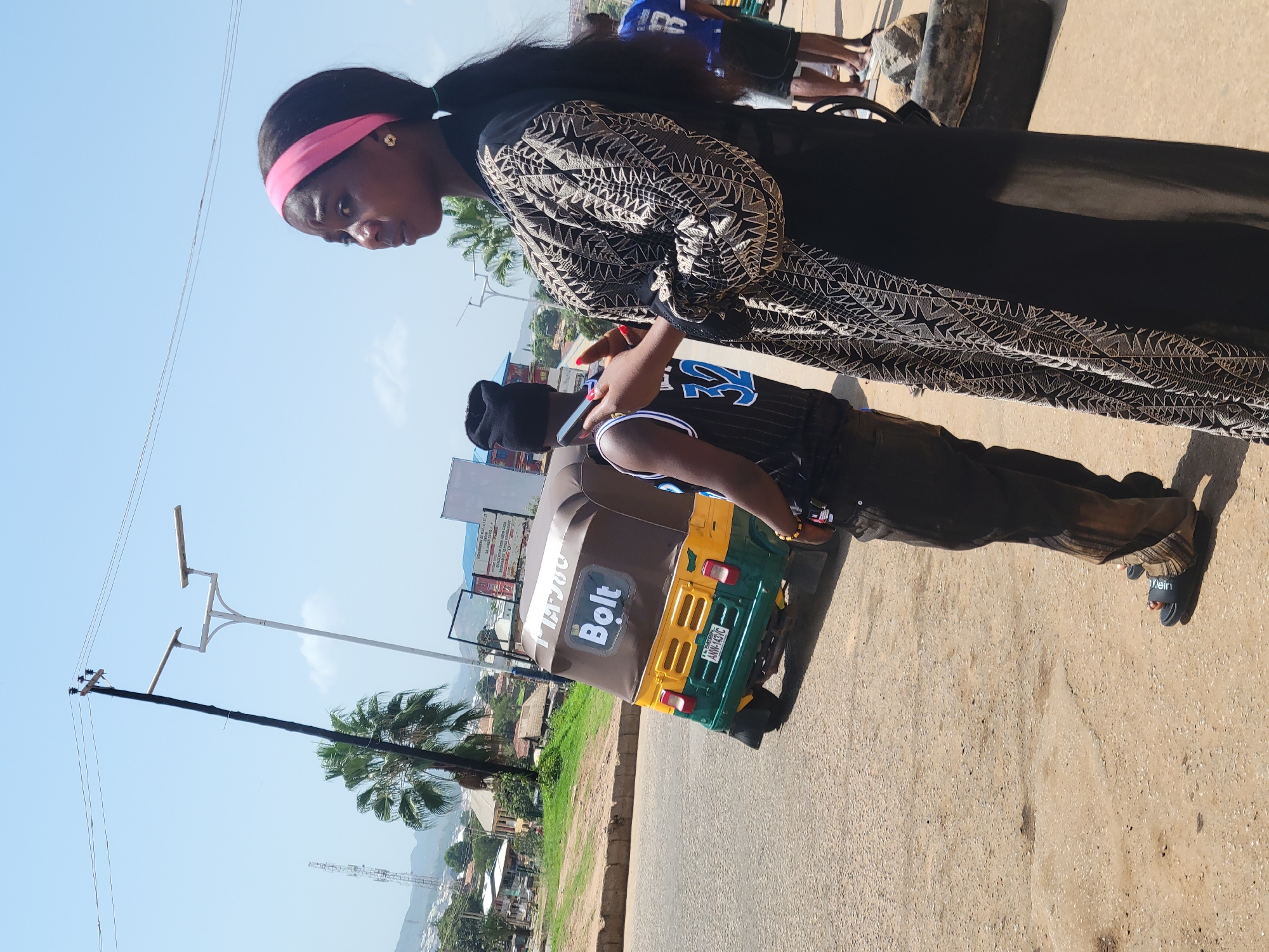 A daytime scene in Rafin Par, Nigeria. A woman with a pink headband is standing on a paved road. She is wearing a black patterned shawl-like garment. A young man wearing a basketball jersey and black beanie is standing next to a Bolt-branded tricycle taxi. In the background, there are buildings, palm trees, and a distant mountain under a clear sky. A black utility pole with streetlights is also visible.