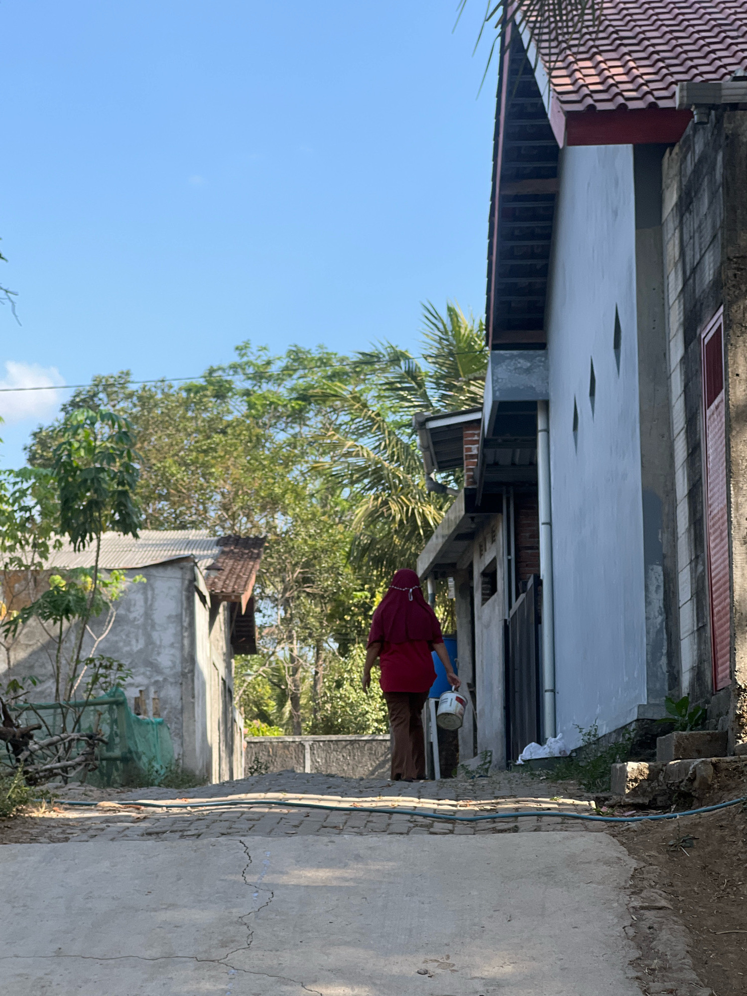In Payaman, Indonesia, a woman wearing a maroon hijab and a maroon shirt walks away from the camera on a paved path lined with buildings. She carries a white bucket with a blue handle in her right hand. The path is cracked and uneven. To the right of the path, a light blue house with a red tiled roof is visible. To the left, a smaller, weathered building with a corrugated metal roof stands behind lush green trees. The sky is a clear, bright blue.