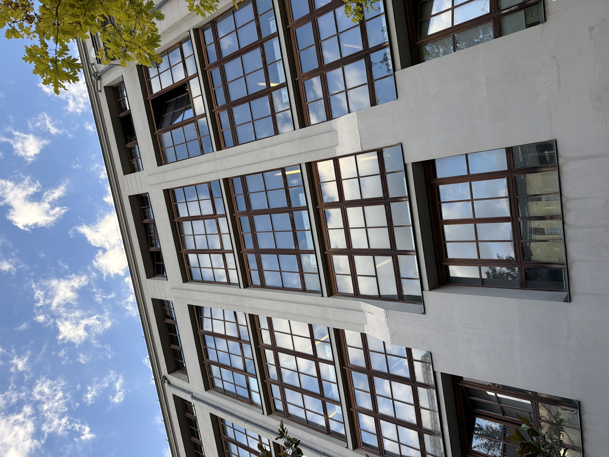 The image shows the exterior of a modern building with large, multi-paned windows with wooden frames. The building is made of a light-colored material, possibly concrete or stucco, and appears to be several stories high. The windows reflect the sky, which is a bright blue with scattered white clouds. There are some green leaves from a tree visible in the upper left corner, suggesting it's a pleasant day, possibly in spring or summer. The architecture of the building, with its repeating window pattern and clean lines, suggests an urban setting, and the provided context indicates this location is Paris, France. There are no people visible in the image. The image is shot from a low angle, looking up at the building, which emphasizes its height and the expanse of the sky. The overall impression is one of modern, clean design and a bright, clear day.