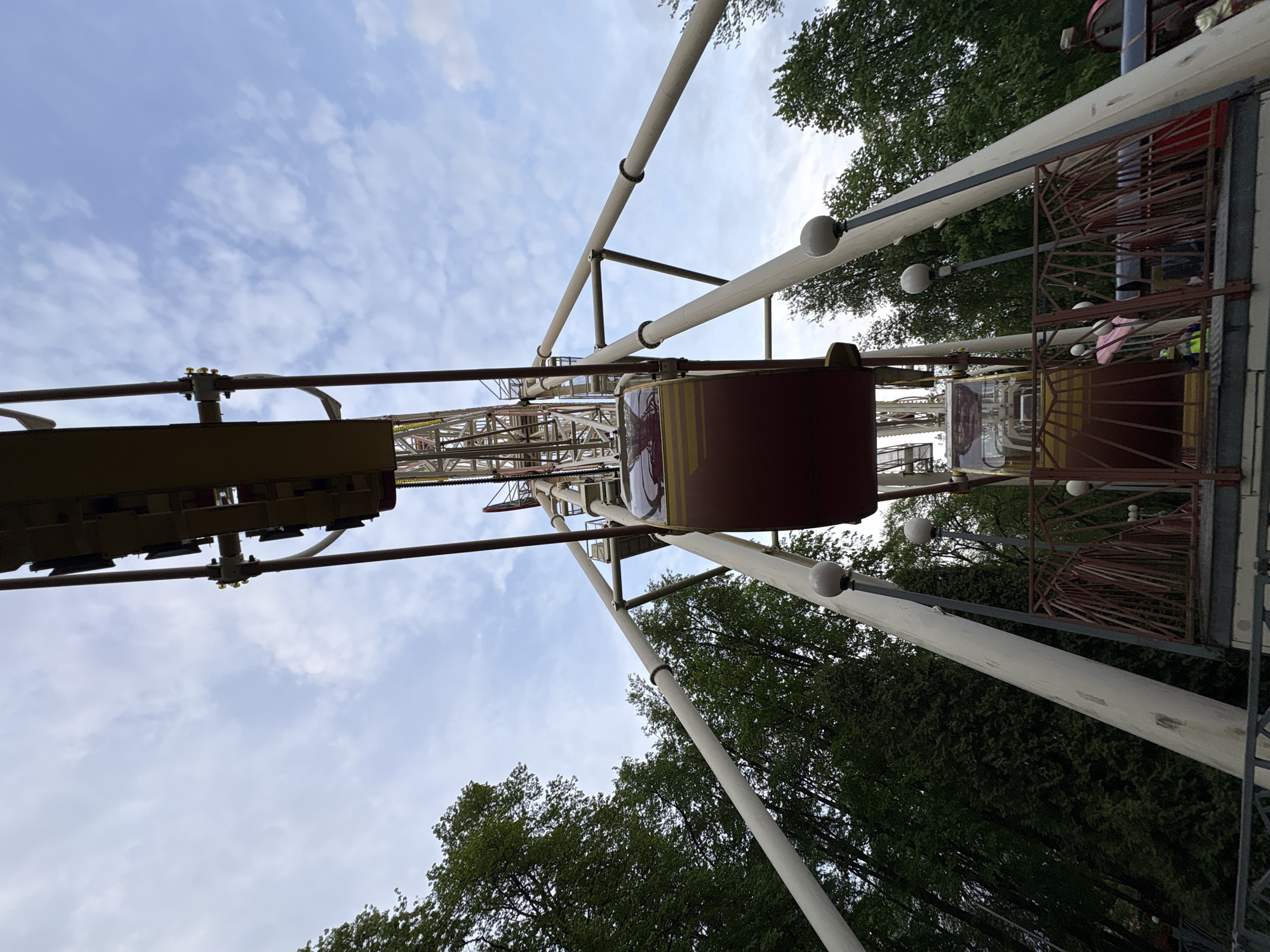 The image shows a partial view of a Ferris wheel in Minsk, Belarus, from a low-angle perspective. The camera is looking upward, capturing the carriages, support beams, and the central structure of the Ferris wheel against a backdrop of trees and a partly cloudy sky. The carriages appear to be empty. The paint is aged and worn.