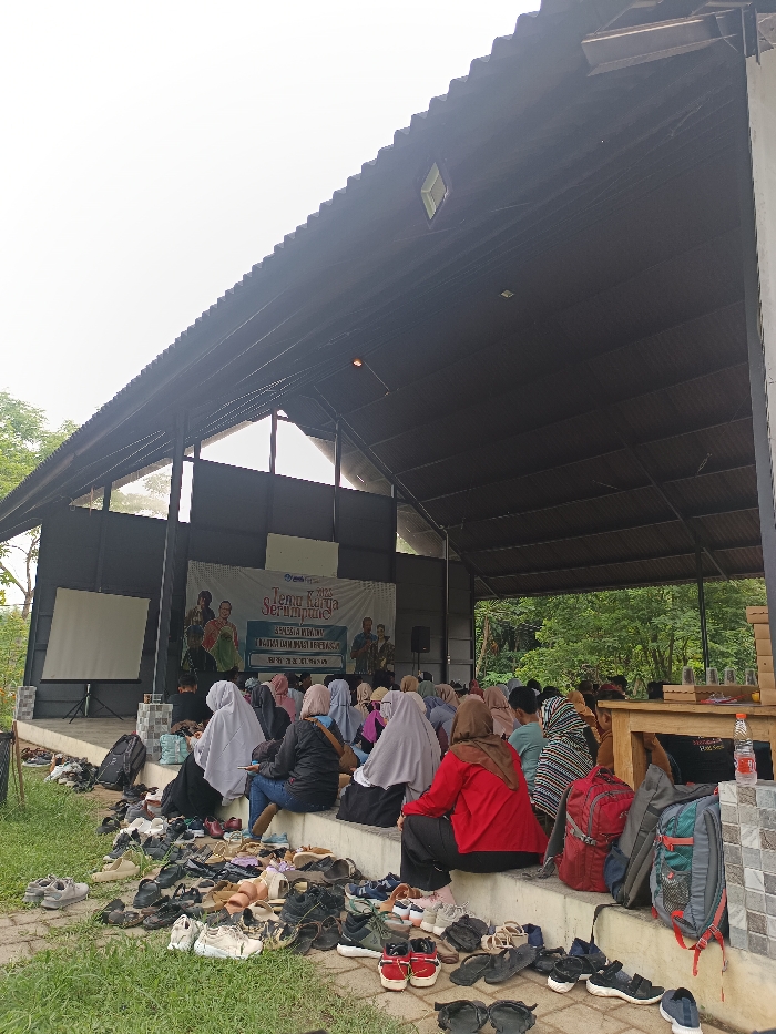 The image displays an outdoor event occurring within a large, open-sided covered structure in Mayang, Indonesia. A dark, corrugated metal roof supported by black metal trusses covers the area. Approximately 30-40 individuals, predominantly women, are seated on a raised concrete platform, facing a stage. Most attendees wear headscarves.
On the stage, a large white banner is displayed, featuring the text "TEMU KARYA SERUMPULE! 2023" along with other Indonesian text and depicted figures. To the left of the banner, a blank white projection screen stands on a tripod. Two male figures are visible on the stage, one holding a microphone, addressing the audience. Speakers are positioned on either side of the banner.
In the foreground, a dense collection of over 50 pairs of shoes, sandals, and sneakers are scattered on a grassy area below the seating platform, indicating removed footwear. Backpacks and personal bags are visible among the seated individuals and next to a wooden table on the far right, which holds several empty glasses and a plastic water bottle. Green trees and dense foliage are visible in the background through the open sides of the structure, under a bright, overcast sky.