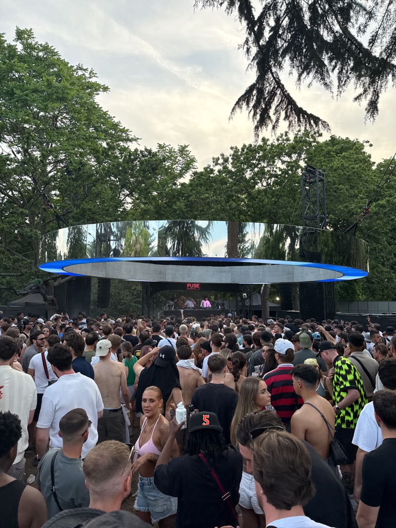 A large crowd of people are gathered in an outdoor space, likely a park or field in Barking, United Kingdom, possibly attending a festival or concert. The crowd is dense, and people are dressed in casual clothing, with some shirtless. Above the crowd is a stage structure with a mirrored circular element and the word "FUSE" visible. Trees surround the area, and the sky is cloudy. A metal structure, probably a tower for lighting or sound equipment, is visible behind the stage. The scene is bustling, with people interacting and enjoying the event.
