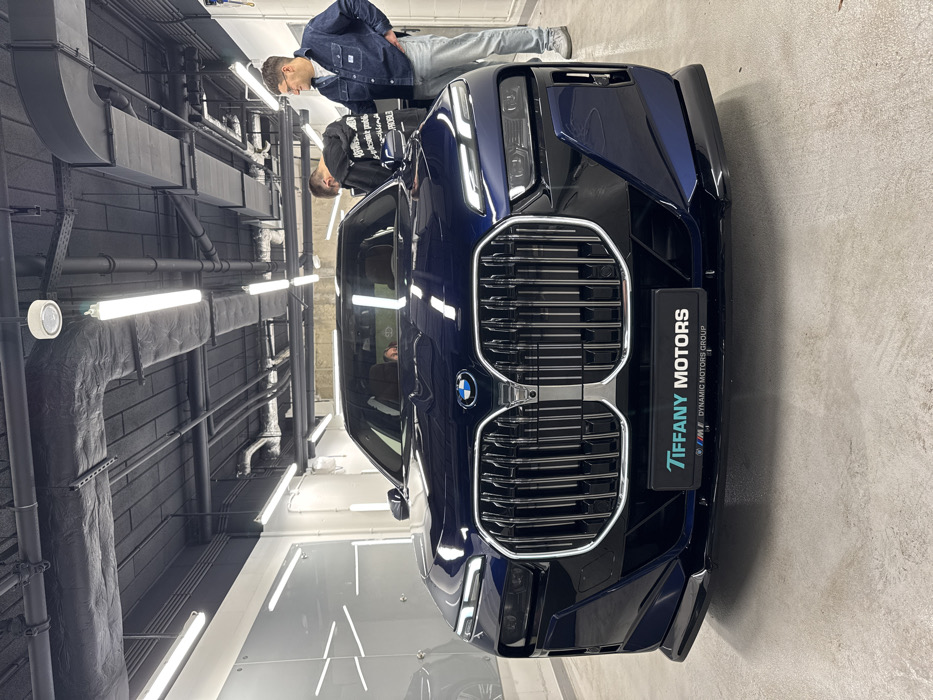 The image depicts the interior of a car detailing facility or showroom belonging to Tiffany Motors, a BMW dealership part of the Dynamic Motors Group, located in Warsaw, Poland.
In the foreground, a dark blue, new-generation BMW 7 Series luxury sedan is positioned centrally, facing the viewer. The vehicle features a prominent, illuminated vertical kidney grille and split headlights. A black license plate frame on the front bumper displays "TIFFANY MOTORS" at the top and "BMW M DYNAMIC MOTORS GROUP" below. The car's surface is highly reflective, indicating a recent cleaning or detailing.
Two adult males are visible behind and to the right of the car. The man on the left wears a black hooded sweatshirt with white text, bending forward and looking down towards the car's hood or windshield, engaged in an action that suggests inspection or adjustment. The man on the right, wearing a blue denim shirt, light-colored jeans, and glasses, stands upright with his left hand on his hip, observing the first man or the vehicle.
The environment features light-colored walls, some with integrated vertical strip lighting and reflective panels on the left. The ceiling is dark grey or black with exposed industrial elements, including ventilation ducts, electrical conduits, and numerous bright fluorescent tube lights, providing ample illumination to the scene. The floor is a light grey concrete or epoxy finish, appearing clean.