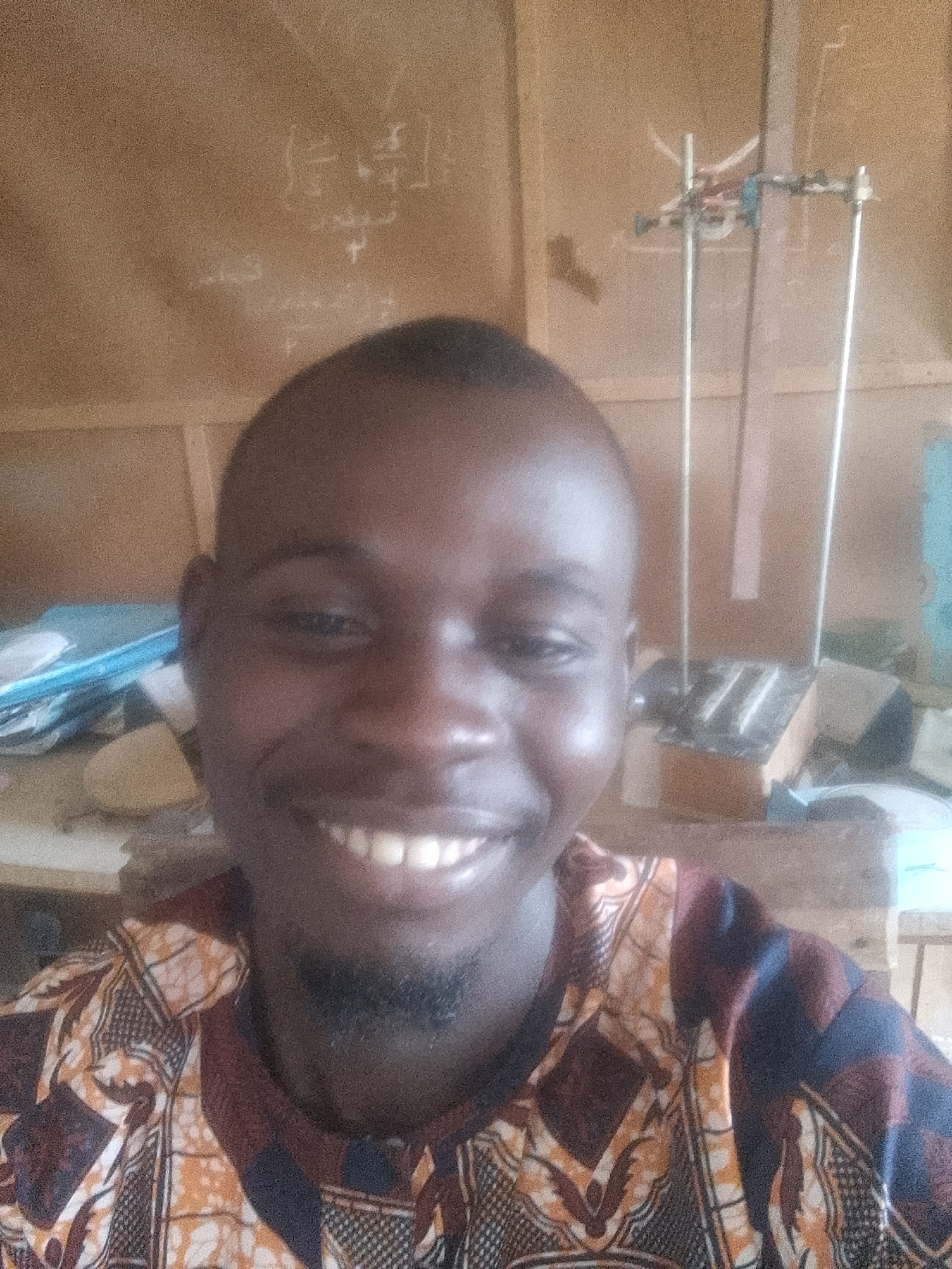 A close-up selfie of a young Black man is captured in a room with wooden walls. He has a shaved head, a faint goatee, and is smiling broadly, revealing his teeth. He is wearing a colorful, patterned shirt with geometric designs in shades of orange, brown, and black. In the background, on the wall behind him, are some faint white markings that appear to be writing or drawings. To his right, a metal stand with various attachments is visible, along with a wooden box. The lighting suggests an indoor environment. The image lacks any specific details that would allow for determination of its location within a city or country.