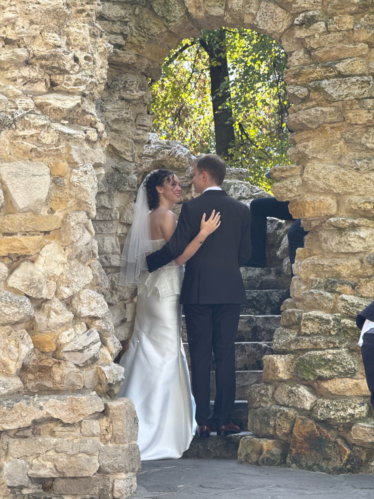 In Moscow, Russia, a wedding couple is captured in an intimate moment within a stone archway. The bride, wearing a white strapless wedding gown and a veil, is embraced by the groom, who is dressed in a black suit and tie. They are standing on stone steps, surrounded by weathered stone structures that suggest an outdoor historical or park setting. Sunlight filters through the leaves of trees visible beyond the archway, creating a dappled lighting effect. The couple is facing away from the camera, looking at each other with smiles, their arms around one another. Their pose and expressions convey affection and connection.