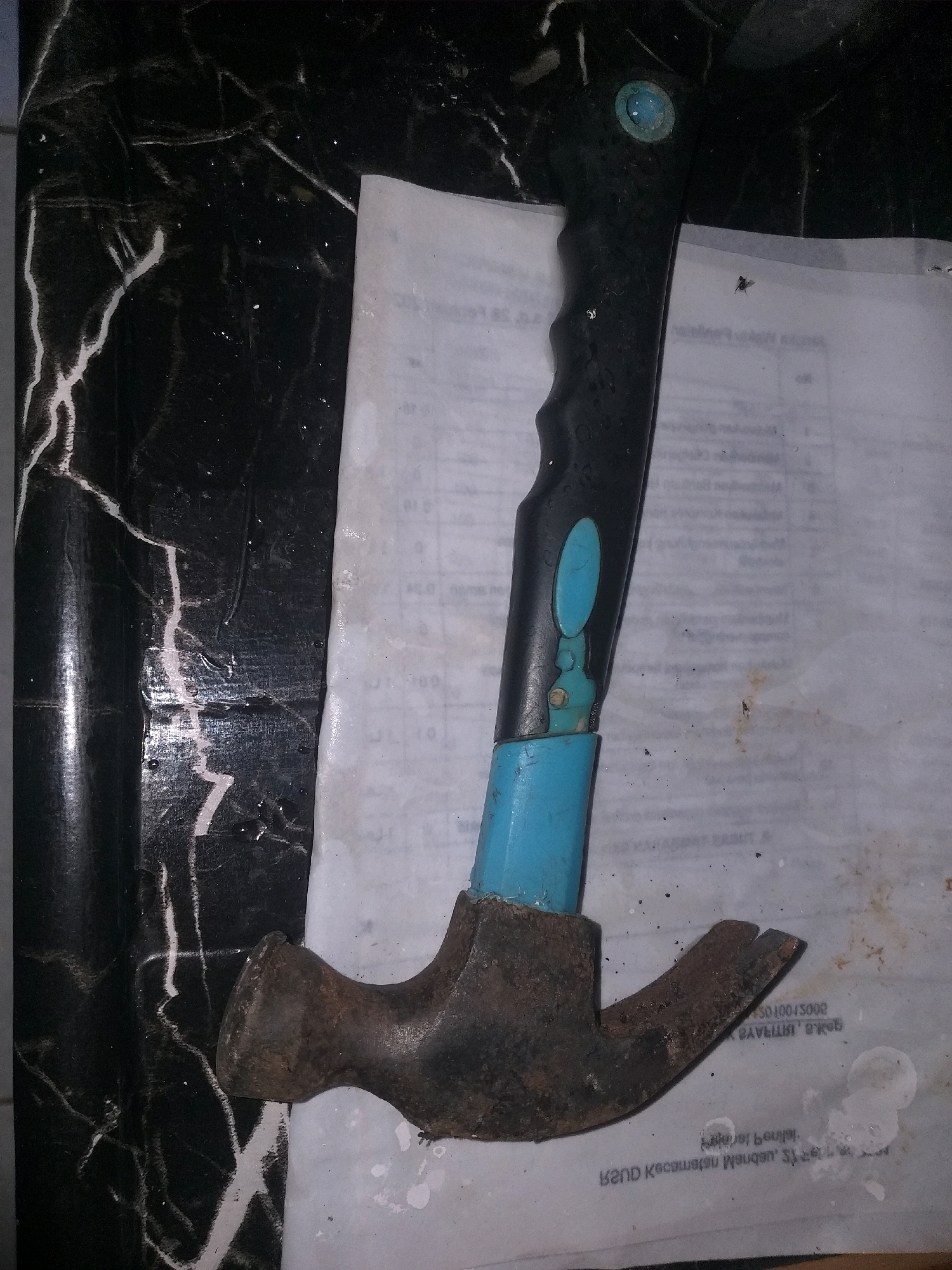 An old, rusty claw hammer with a black handle and blue accents rests on top of a piece of paper. The paper is lying on a black surface with white marbled streaks, possibly a countertop. The background is dark and out of focus, with some specks of light. The image was taken in Air Jamban, Indonesia.