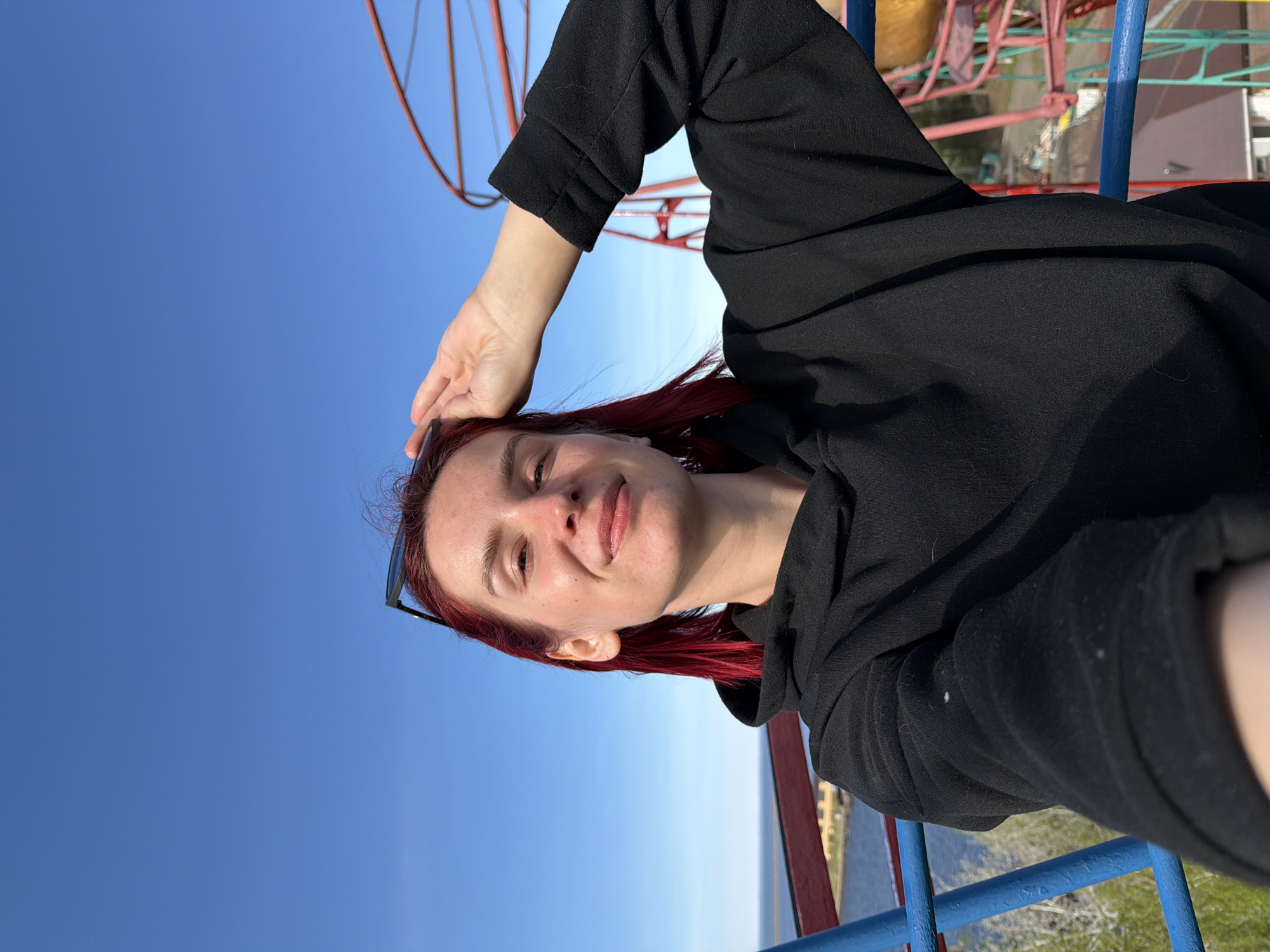 A woman with red hair, wearing a black hooded sweatshirt, is in the city of Chaikovsky, Russia. She is on a Ferris wheel. A section of a metal railing is in the foreground. The sky is blue. In the background is foliage and buildings.