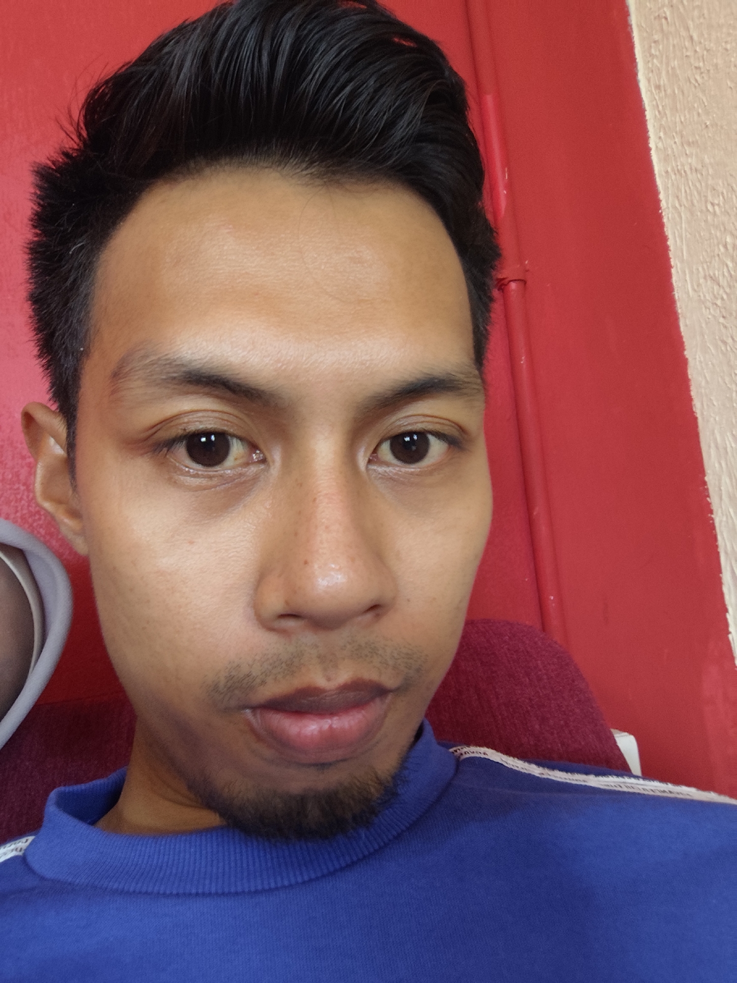 The media file is a close-up, waist-up selfie of a man. He has dark hair styled upwards, a prominent forehead, dark eyes with light brown irises, a light beard, and dark lips. He is wearing a royal blue crew-neck sweatshirt with white stripes on the shoulders. The background is a vibrant red, with a textured beige surface visible on the right side. The lighting appears to be artificial, possibly from an indoor setting, and there is no indication of the time of day or weather conditions. The man's expression is neutral, and he is looking directly at the camera. The overall scene is simple and focused on the subject. There is no visible text in the image.