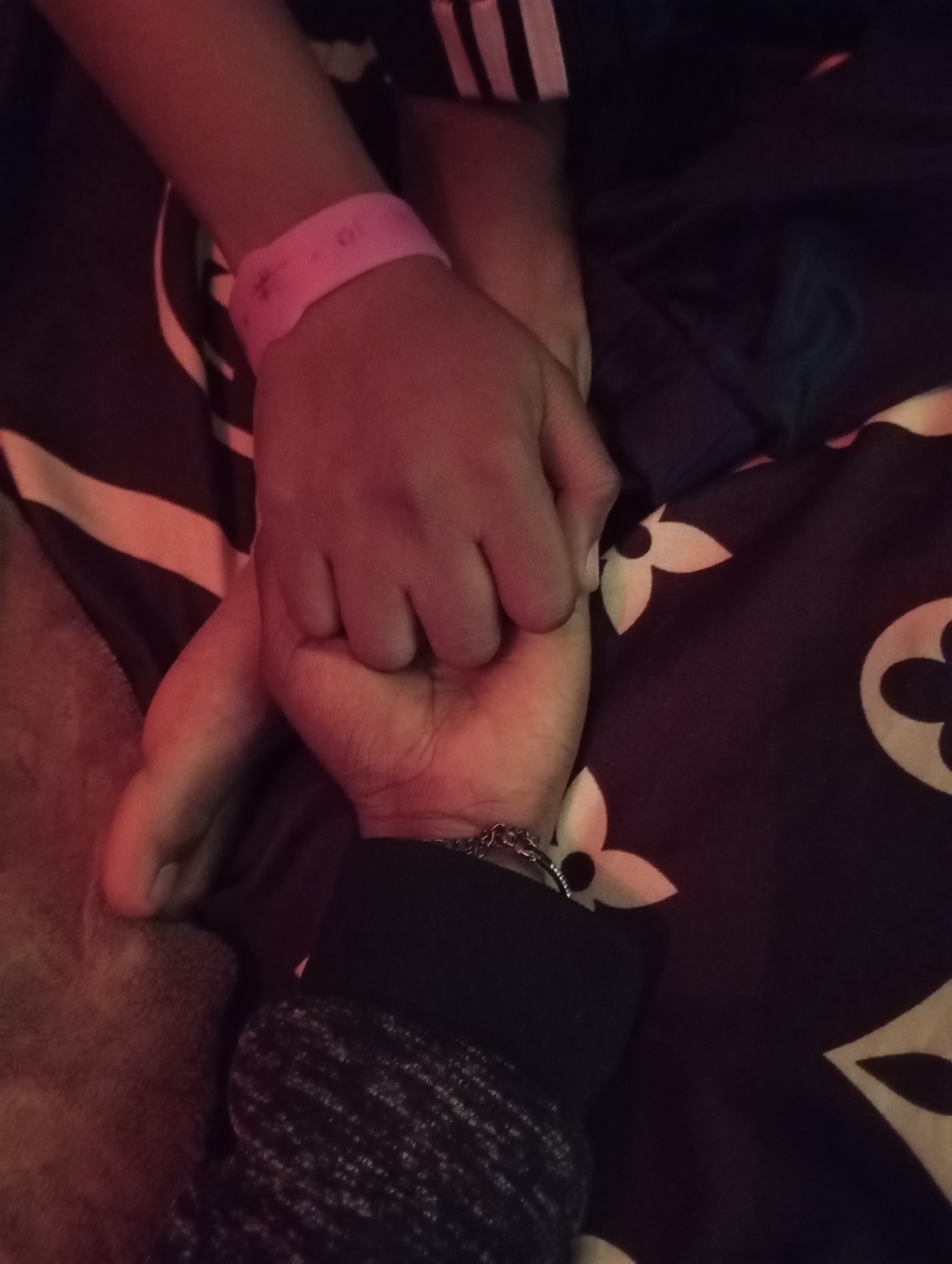 The image shows a close-up of two hands clasped together. The hand on the left belongs to a child and has a pink wristband with a small imprint of a flower. The hand on the right belongs to an adult and is wearing a bracelet with silver chain links. Both hands are resting on a dark fabric with a pattern of stylized flowers. The lighting suggests an indoor setting, possibly at night. There are no discernible landmarks or contextual clues to determine the specific location within Jos, Nigeria. The scene is intimate and appears to depict a moment of connection or comfort between a child and an adult.