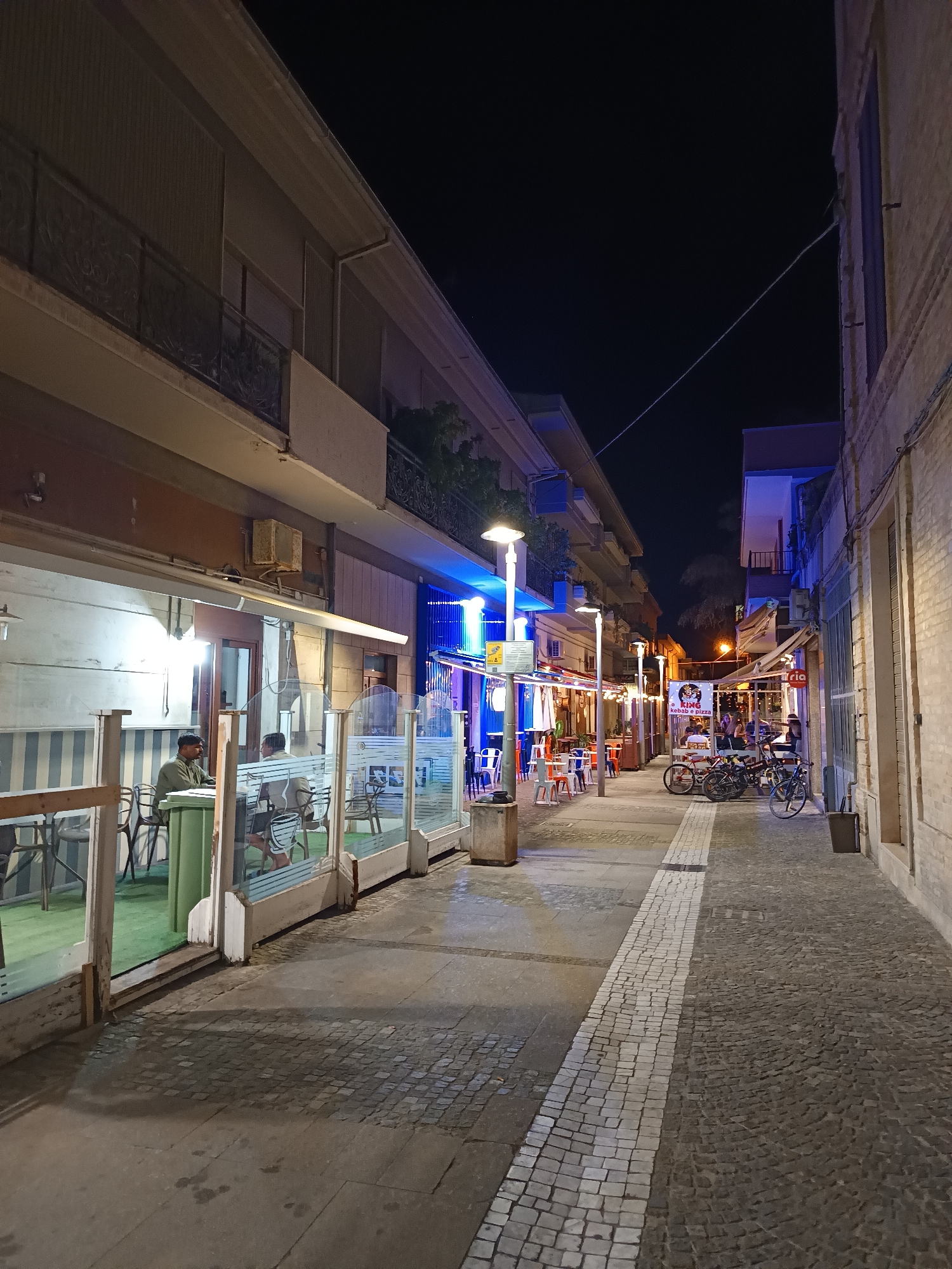 The image depicts a nighttime street scene in Roseto degli Abruzzi, Italy, likely a commercial area. The perspective is from a low angle, looking down a pedestrian walkway lined with buildings on either side. On the left, a storefront with outdoor seating is visible, featuring glass partitions, tables, and chairs. Two men are seated at a table inside the partially enclosed area. The building on the left has a second story with a balcony and ornate railings.
The walkway itself is paved with cobblestones and features a distinct white stripe dividing the path. Streetlights illuminate the scene, casting a warm glow. Several bicycles are parked on the right side of the walkway. Further down the street, more storefronts with outdoor seating, colorful lights, and signs are visible, suggesting a lively atmosphere, possibly restaurants or cafes. The overall impression is of a quaint, European street at night, with activity spilling out from the establishments onto the pedestrian area. The presence of outdoor seating and bicycles suggests a walkable and potentially tourist-friendly environment.