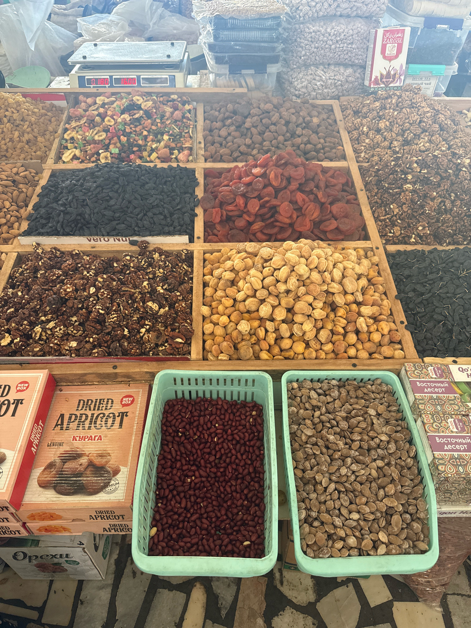 This is a high-angle, medium shot of a bustling market stall in Tashkent, Uzbekistan, specializing in dried fruits and nuts. The stall is overflowing with various edible items displayed in wooden crates and plastic baskets. In the foreground, to the left, are boxes of "Dried Apricot" and "Opexu" (likely a brand or type of nut/fruit). Next to these boxes are two light green plastic baskets filled with dried goods: the left basket contains dark red, small, oval-shaped items (likely beans or small dried fruits), and the right basket holds light brown, flat, oval seeds.
Behind these, a variety of nuts and dried fruits are meticulously arranged in wooden compartments. These include piles of dark, small raisins, light brown dried apricots, and a large quantity of chopped walnuts mixed with white nut pieces. Further back, on a scale, are bags of various nuts and dried fruits. Some bags are translucent, allowing glimpses of their contents, while others are opaque. A box with Persian writing, labeled "ZARGOL," is visible in the upper right section, suggesting the market's international influences. The overall environment appears to be an outdoor or semi-outdoor market, with rough flooring and a general sense of abundance and commerce. The lighting is bright, suggesting daytime. No people are directly visible in the frame, but the arrangement and variety of goods imply active trade.