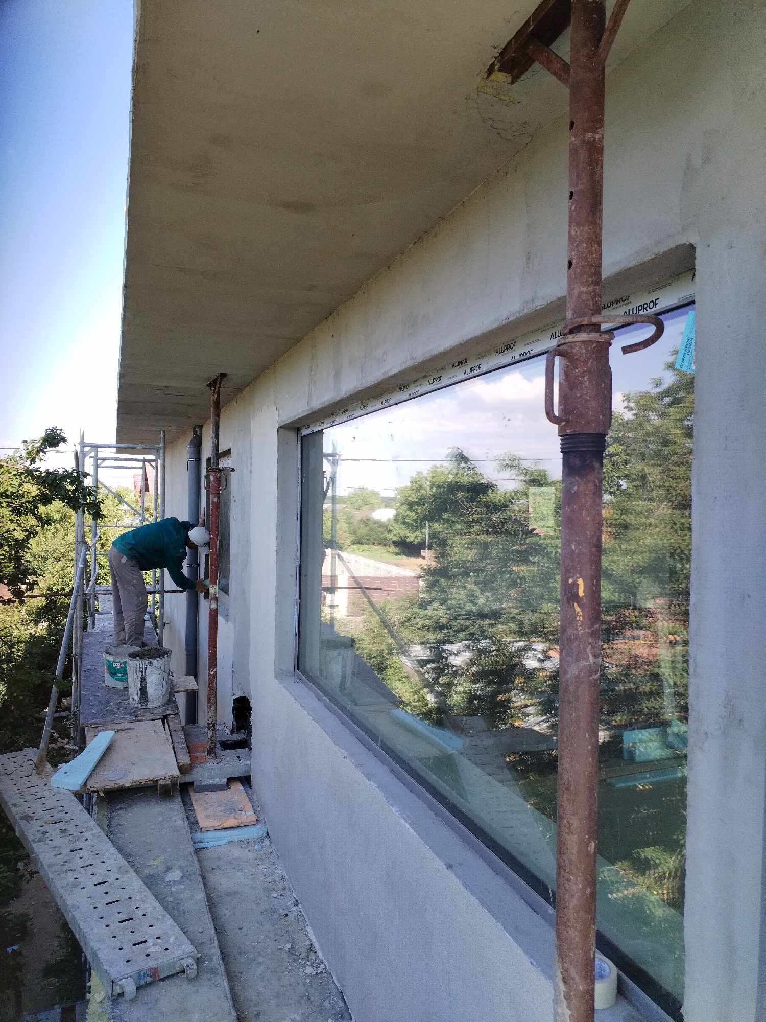 In Dumbrăveni, Romania, a construction worker is seen on scaffolding, applying stucco to the exterior of a building. The worker, dressed in a teal jacket and beige pants with a white hard hat, is actively engaged in the stucco application process near a large, reflective window. The window offers a view of lush green trees and the sky. The construction site is characterized by concrete structures, scaffolding, and various building materials, including buckets of what appears to be stucco. The overall scene depicts an active construction phase with clear evidence of manual labor and building progress.