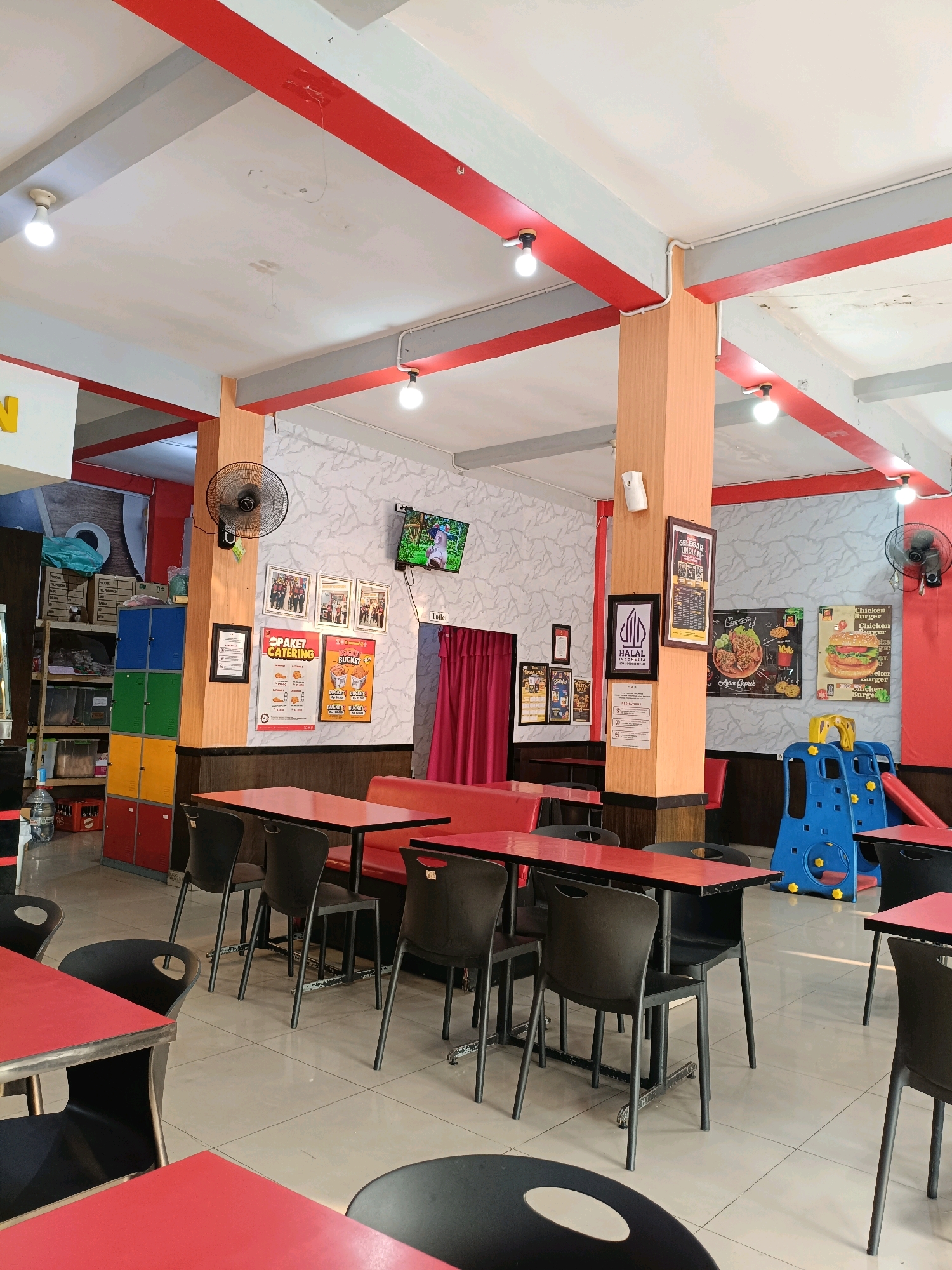 The image depicts the interior of a restaurant in Ngemplak, Indonesia. The restaurant features a red and white color scheme with red beams on the ceiling and red tabletops. Various tables and chairs are arranged throughout the space, with some tables set with red tops and black chairs, while others have red bench seating. The walls are decorated with patterned wallpaper and framed pictures and posters, including advertisements for catering, a burger, and a "Halal Indonesia" certification.
In the background, there are shelves stocked with boxes and containers, as well as a stack of colorful lockers. A small children's play area with a blue slide and yellow steps is visible on the right side of the image. Two wall-mounted fans are present, one on a central wooden pillar and another on the right wall. A television screen is mounted high on the wall on the left side, displaying an image. A sign indicates "Toilet" with a curtained doorway nearby. Natural light filters in from the side, and overhead lighting fixtures are also visible. No people are visible in the image.