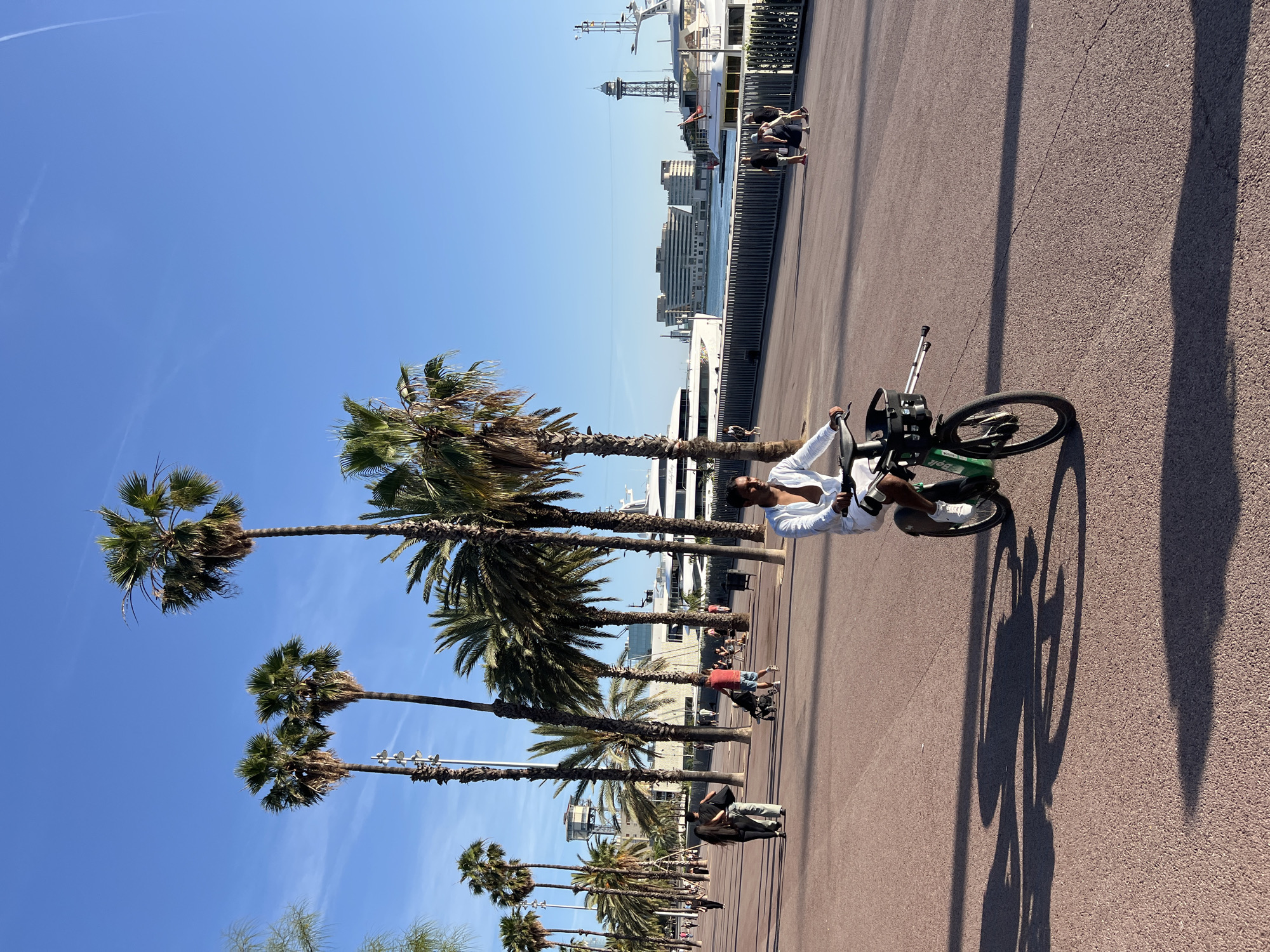 The image depicts a wide, paved promenade alongside a harbor in Barcelona, Spain, likely within the Port Vell area, with the World Trade Center Barcelona building visible in the background. The scene is illuminated by strong sunlight, casting long shadows.
In the foreground and slightly to the right, a man with dark skin, dark hair, and sunglasses is riding a green and black electric bicycle branded "Zolti." He wears a white long-sleeved shirt, unbuttoned, white shorts, and white sneakers. He is positioned on the bicycle with his right foot on the pedal and his left leg extended slightly forward, and he appears to be looking towards his right. The bicycle features a front basket containing two silver cylindrical objects extending horizontally forward, resembling poles. A distinct shadow of the man and the bicycle is cast diagonally across the reddish-brown asphalt surface of the promenade.
The promenade is wide and appears to be a pedestrian and cycling path. Numerous palm trees, varying in height, line the left side of the promenade, casting long shadows to the left. Several people are visible in the mid-ground: a couple walks away from the camera, the woman in black and the man in light-colored attire; further back, a person in a red shirt pushes a baby stroller. Scattered groups of people are visible further down the promenade and near the water's edge.
To the right, a dark metal fence or railing separates the promenade from the water of the harbor. Several large, white yachts are docked along the waterfront. One yacht, visible on the left side of the harbor, clearly shows "Lady Lola" written on its stern. In the background, across the water, is a large, multi-story building with a distinctive curved, modern architectural style (the World Trade Center Barcelona). To the right of this building, a tall, slender, lattice-work tower stands prominently against the horizon. The water is calm and reflects the clear blue sky. The sky above is a uniform clear blue with a few faint, wispy contrails visible at high altitude.