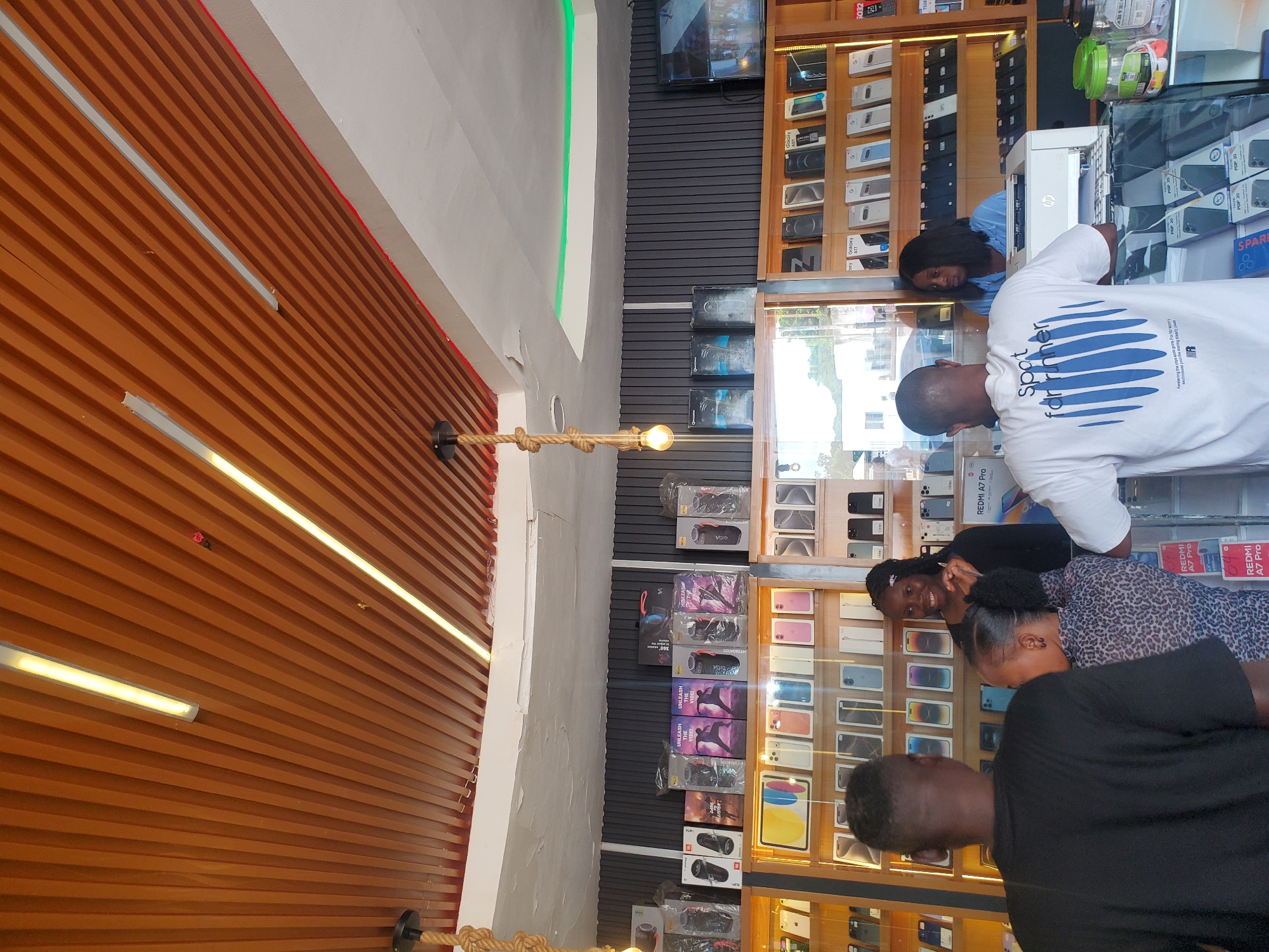 The image depicts the interior of a cell phone shop, likely located in Jos, Nigeria, as indicated by the style of the shop and its offerings. The shop features a modern design with a slatted wooden ceiling and fluorescent lighting. Various brands of cell phones and accessories are displayed on shelves behind glass counters. In the foreground, three individuals are visible. Two men are standing, one with his back to the camera wearing a black t-shirt, and another partially visible on the right wearing a white t-shirt. A woman with dark hair is standing behind a counter, interacting with a customer who is not fully visible. The shelves are stocked with numerous cell phone boxes, with brands like Samsung and Apple appearing to be prominent. A printer is situated on the counter. The overall atmosphere suggests a retail environment focused on electronics sales.