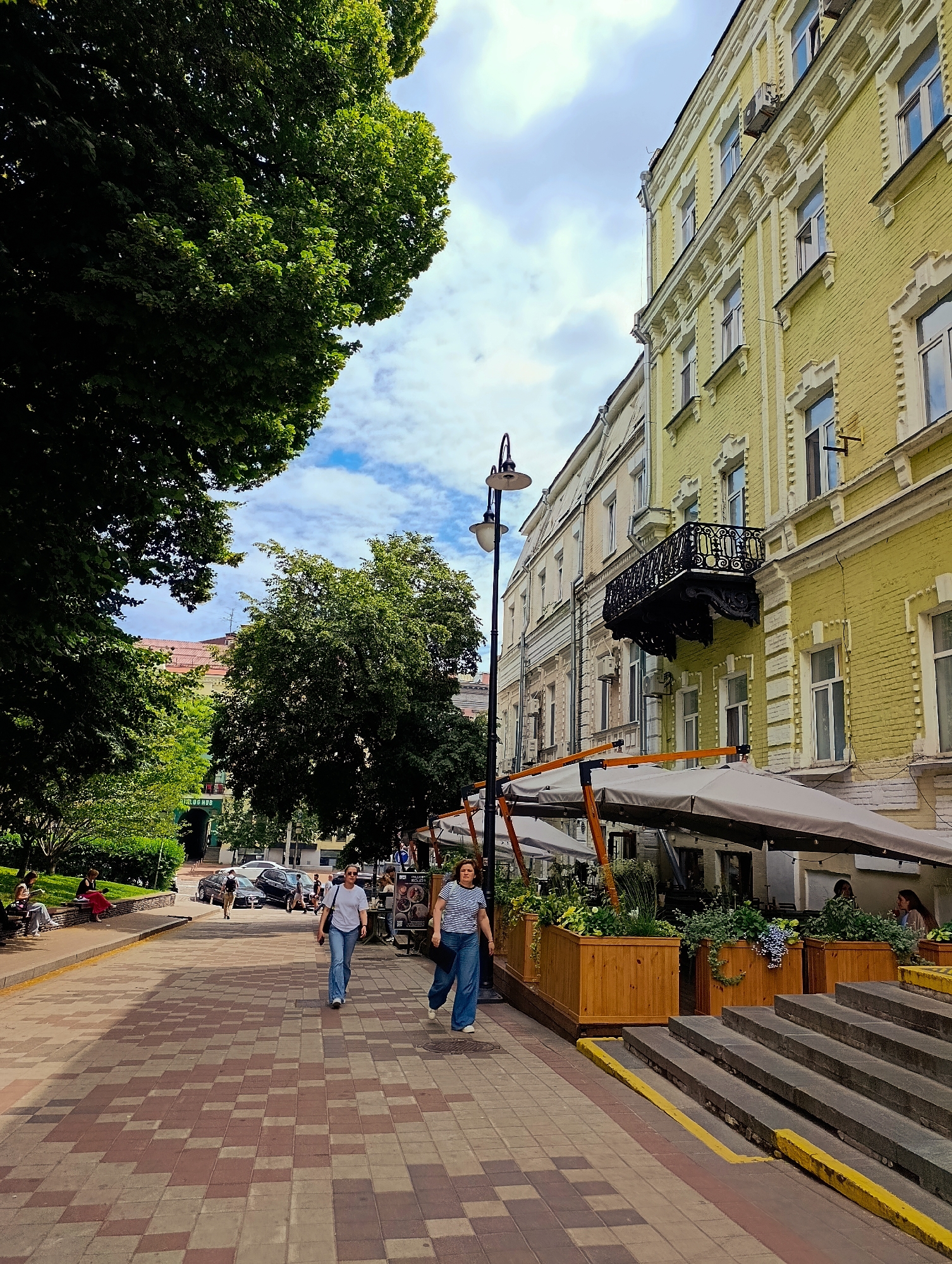 This is a vertical, eye-level, medium shot of a street scene in Kyiv, Ukraine, on a cloudy day. The perspective looks down a brick-paved walkway lined with trees on the left and a yellow building with decorative architecture on the right. Two women are walking towards the camera on the walkway. The woman on the left wears a white t-shirt and blue jeans, and the woman on the right wears a striped t-shirt and blue jeans. Several people are seated on a bench in the background on the left, under the trees. Cars are parked further down the street. Outdoor seating for a restaurant with umbrellas and planters is visible on the right, extending from the yellow building. A lamppost stands near the left edge of the walkway.