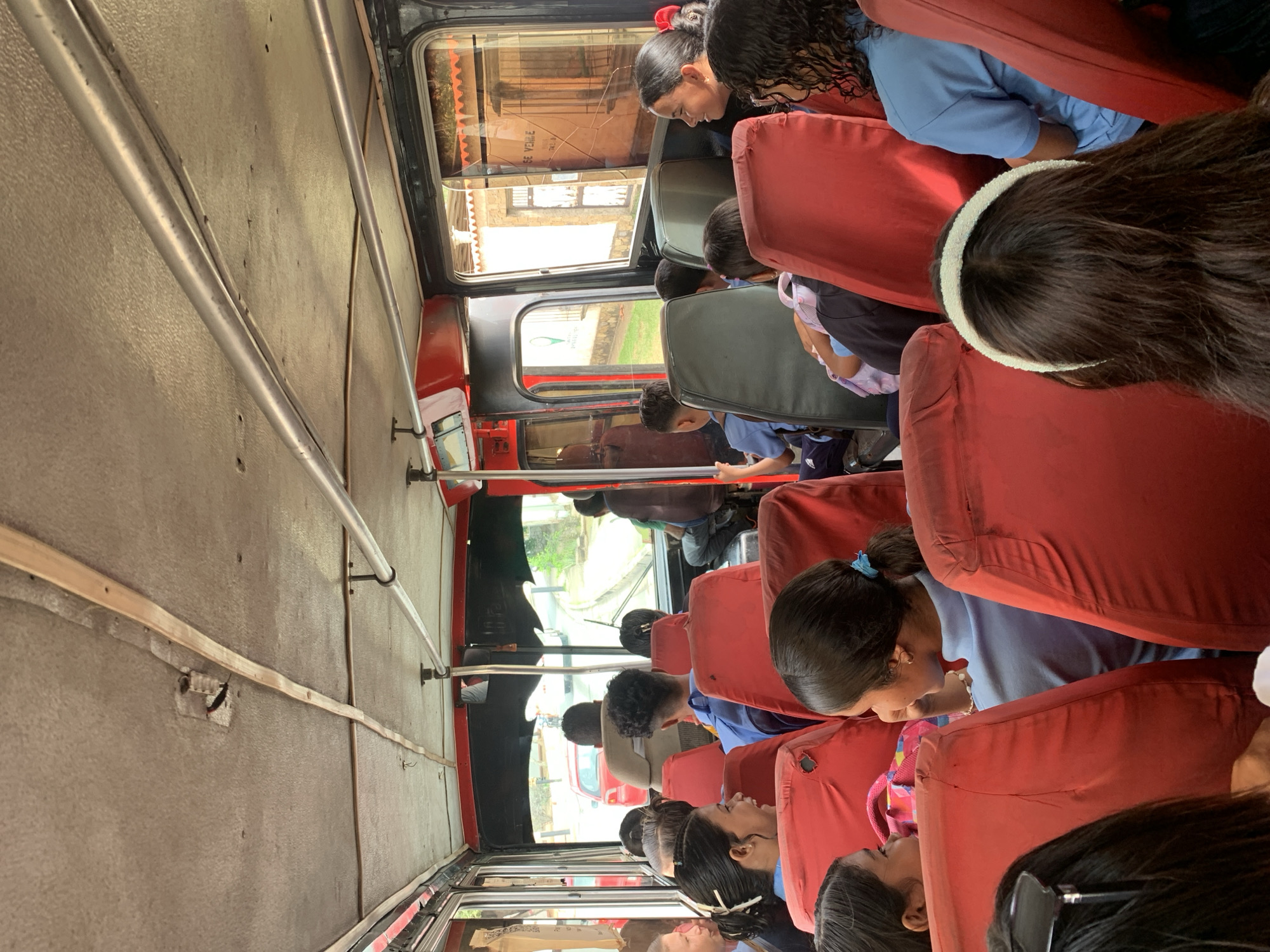 The image depicts the interior of a red bus filled with passengers, predominantly children wearing blue uniforms, suggesting a school bus. The bus appears to be in transit, as indicated by the view through the windows of a street with passing vehicles and buildings. One building visible outside has a sign that reads "SE VENDE," which translates to "FOR SALE" in Spanish. The bus interior features rows of red seats, overhead metal handrails, and a textured ceiling. The passengers are mostly facing forward, with some engaged in conversation. The overall scene suggests a typical day on a school bus in an urban or semi-urban environment in La Tagua, Venezuela.