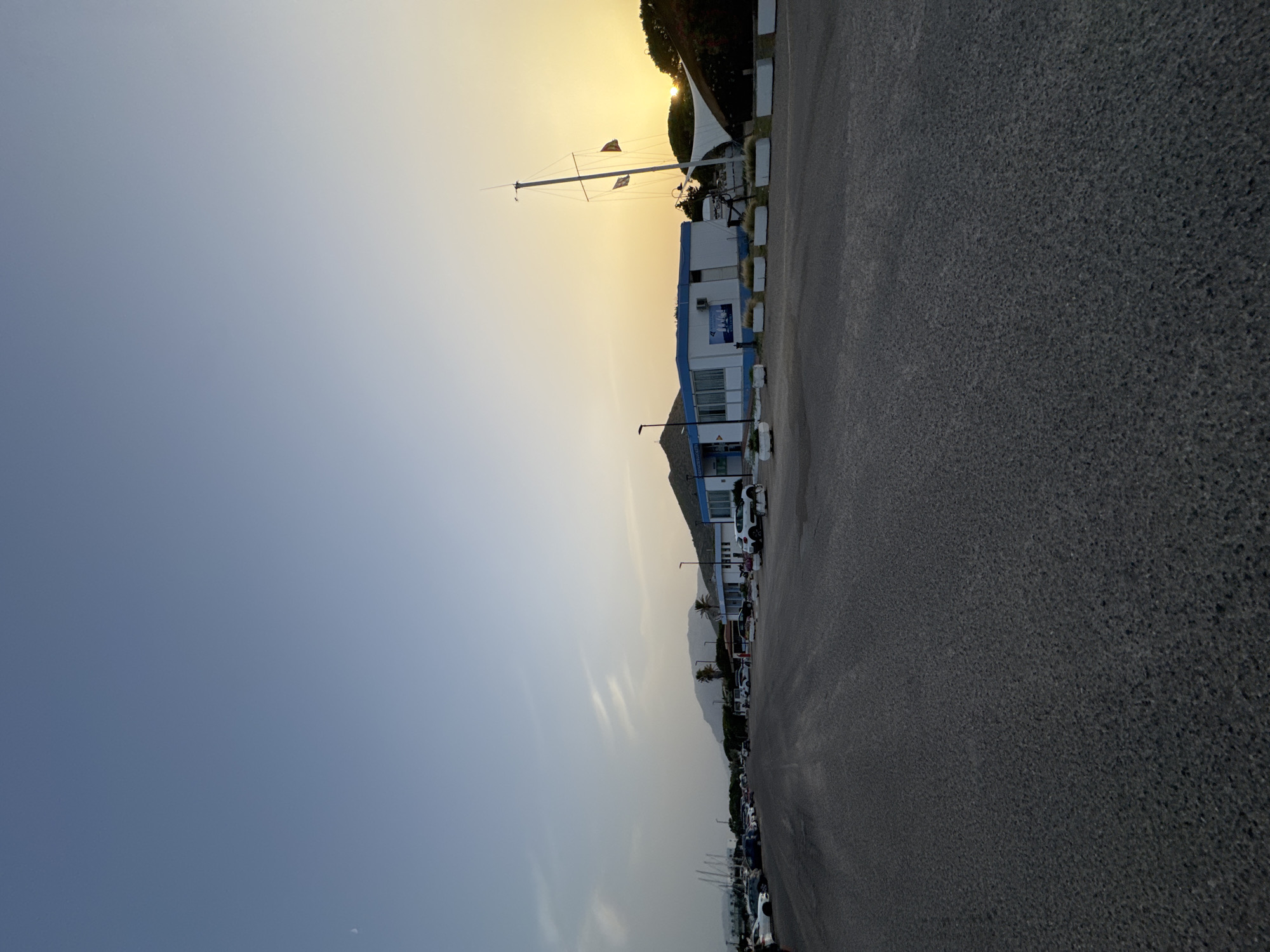The image captures a scene at dusk in San Giovanni, Italy, likely near a marina or coastal area. In the foreground, a wide asphalt road or parking lot stretches towards the horizon. To the right of the road, a low-lying building with a white facade and blue trim is visible. In front of this building, there are several white planters with some vegetation. Adjacent to the building is a large, white, sail-like structure supported by a mast, resembling a boat's sail. Two flags are attached to the mast. The sun is low on the horizon, casting a warm, golden glow and creating a sunset effect. In the background, hills or mountains are silhouetted against the sky. A line of parked cars is visible along the left side of the road. The sky transitions from a clear blue at the top to a soft yellow and orange near the sunset.