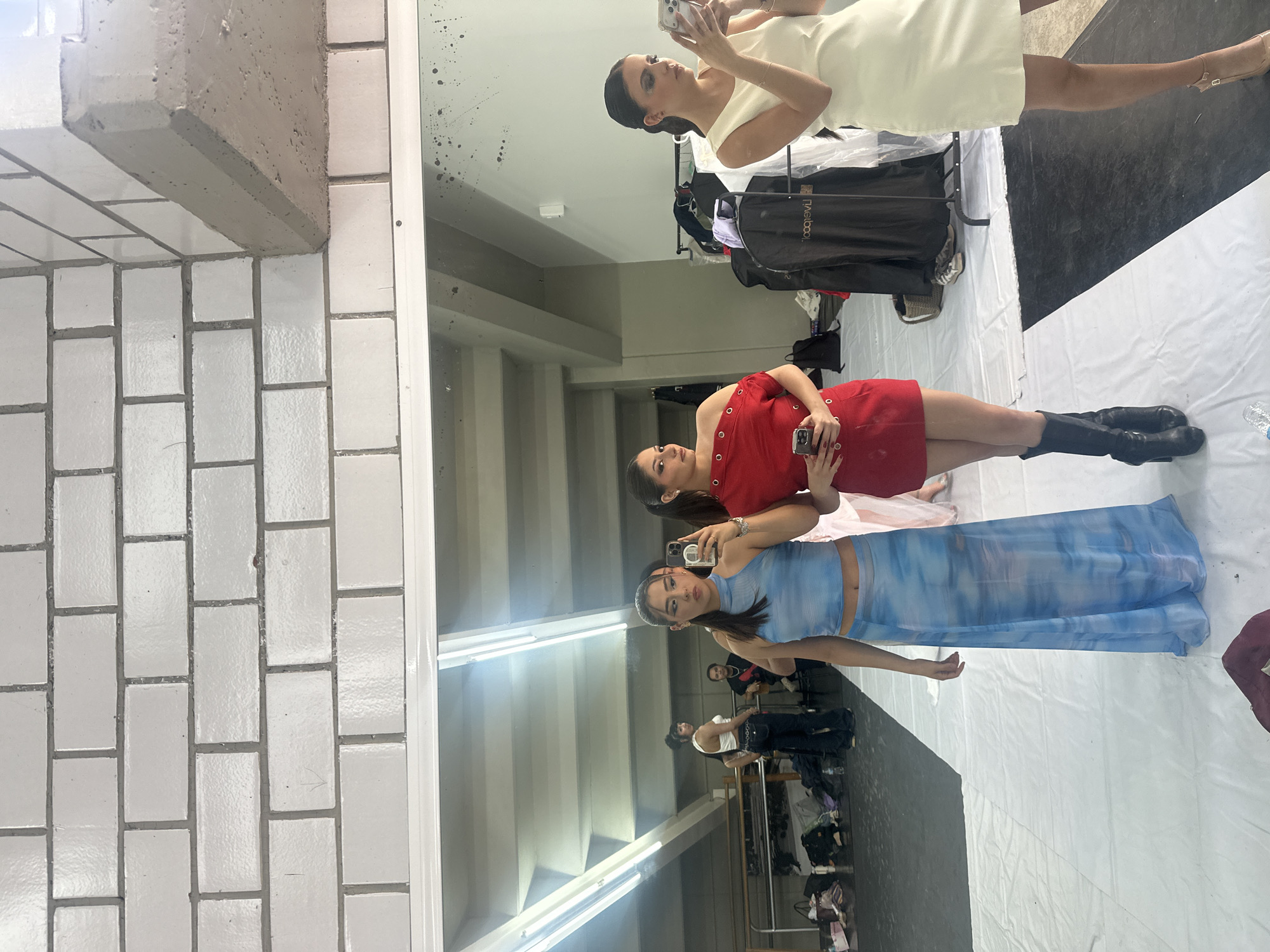 In an indoor setting in San Bernardino Tlaxcalancingo, Mexico, a large mirror reflects three women and the surrounding space. The image is oriented with a white tiled wall and concrete beam on the left, framing the mirror that fills the majority of the frame.
In the mirror's reflection, three women are visible, seemingly taking selfies or interacting with their phones. The woman closest to the top of the frame wears a cream or off-white sleeveless dress and is holding a phone, angling it to capture her reflection. Below her, a woman in a red, one-shoulder dress with eyelet details also holds a phone up, looking into it. Further down, a third woman wears a sheer, light blue patterned two-piece outfit consisting of a crop top and a long skirt, and she too is holding a phone.
The floor in the reflection is a mix of black and white, possibly white tarps laid over a dark floor. Various items are visible in the background reflection: a black clothing rack with garment bags, one of which clearly displays "naelboo" in white text, and a pair of white sneakers on the floor. A woman in a white top and dark pants is faintly visible in the deeper background, standing near a ballet barre. The mirror itself has notable smudges and dark splatters, particularly in the upper left corner of the reflected area. The lighting appears bright, suggesting a well-lit studio or changing room environment during the day. The overall scene suggests a moment of preparation or a break in a fashion-related or performance setting.