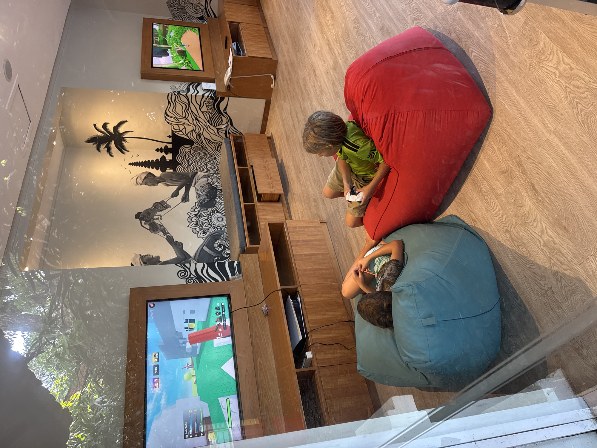 A photo depicts two children playing video games in an indoor entertainment area. One child is seated on a red beanbag chair, holding a game controller, while the other is on a blue beanbag. In the background, there are two wall-mounted televisions displaying game visuals. The interior design includes wooden shelving and wall murals, and is likely located in the city of Kuta Selatan, Indonesia.