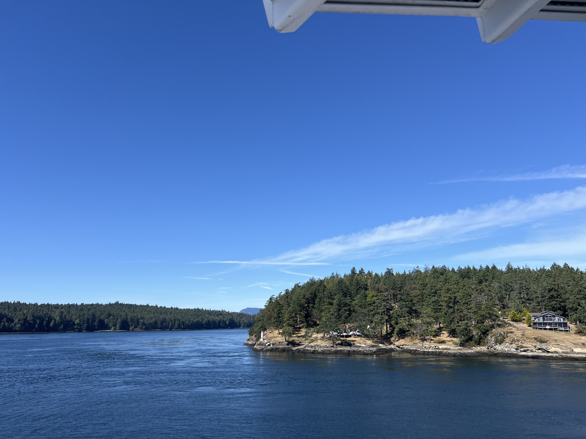 A wide shot captures a body of water with a dense, evergreen forest on the right bank. The water is deep blue and calm, with gentle ripples. A prominent peninsula juts out into the water, covered with trees and dotted with a few houses. A white lighthouse stands on a rocky outcrop at the tip of the peninsula. In the distance, another forested shoreline is visible. The sky is a clear, vibrant blue with a few wispy white clouds. A portion of a white structure, possibly part of a boat or building, is visible at the top of the frame.