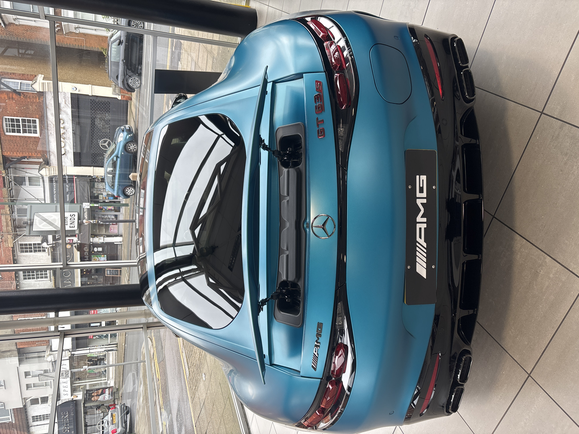 Here's a description of the image:
The image shows the rear of a matte blue Mercedes-AMG GT 63 S car. The car is inside a dealership or showroom with a large glass window. Through the window, a street scene is visible in a town in the UK. The reflections in the car's rear window mirror the urban landscape. The car's badges and details, such as the "AMG" logo and "GT 63 S" designation, are visible. The car is parked on a tiled floor.